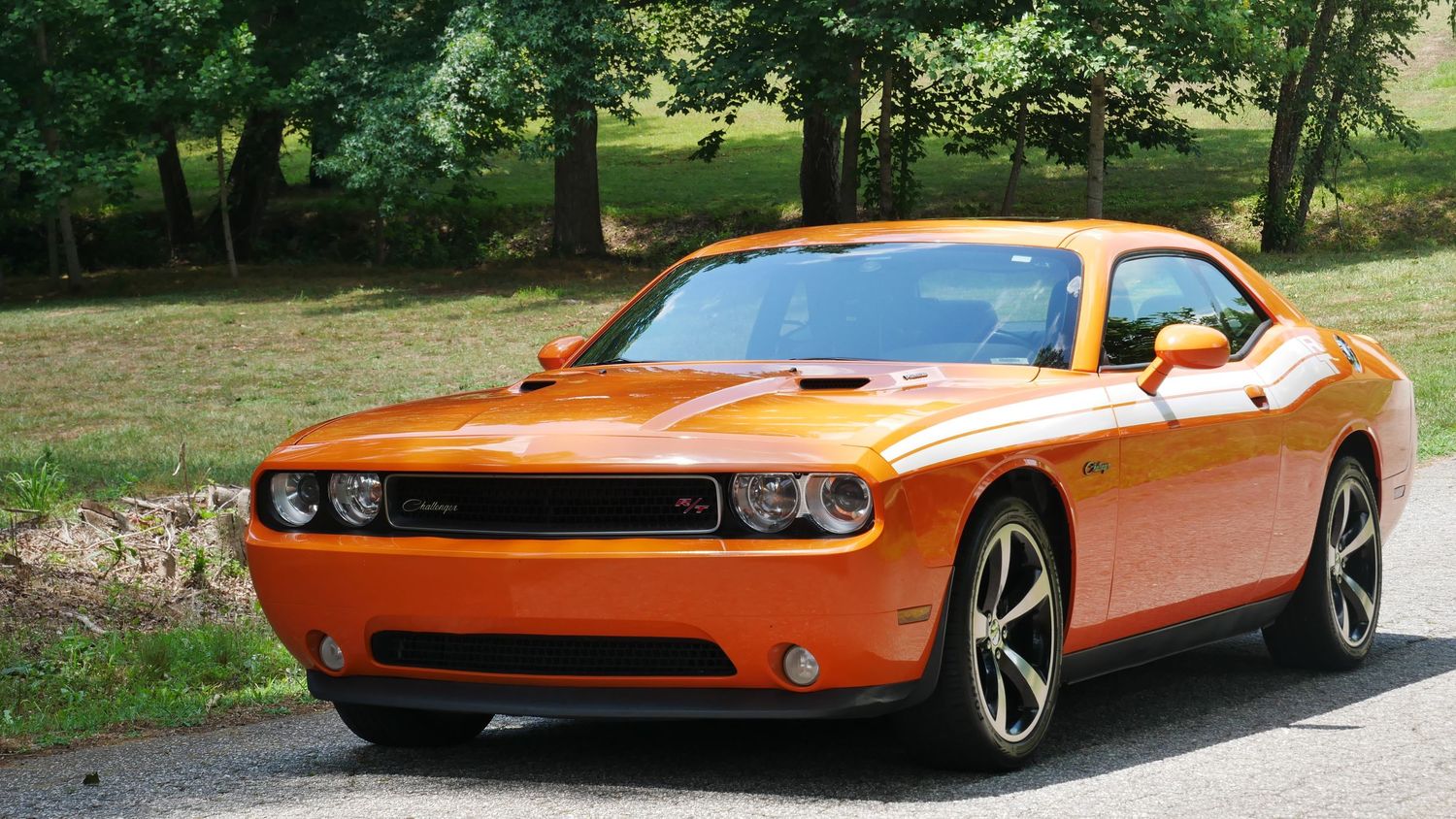 An orange Dodge Challenger driving along a tree-lined road with grassy lawns on either side.
