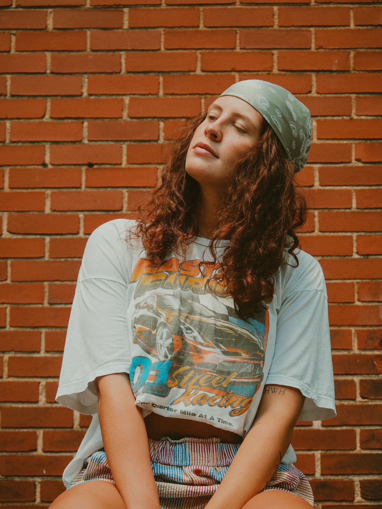 A person with curly hair sits casually against a red brick wall wearing a light t-shirt.