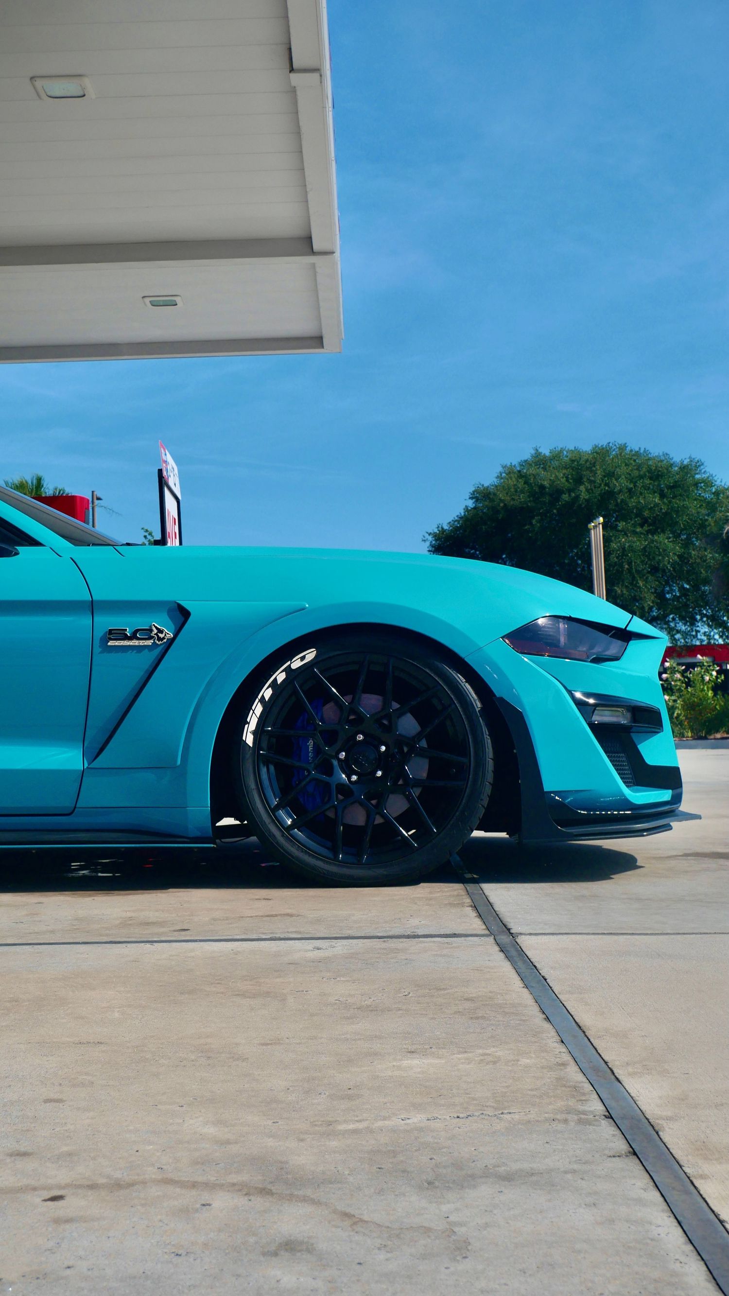 Close-up of a turquoise Mustang's front wheel and side panel against a blue sky.