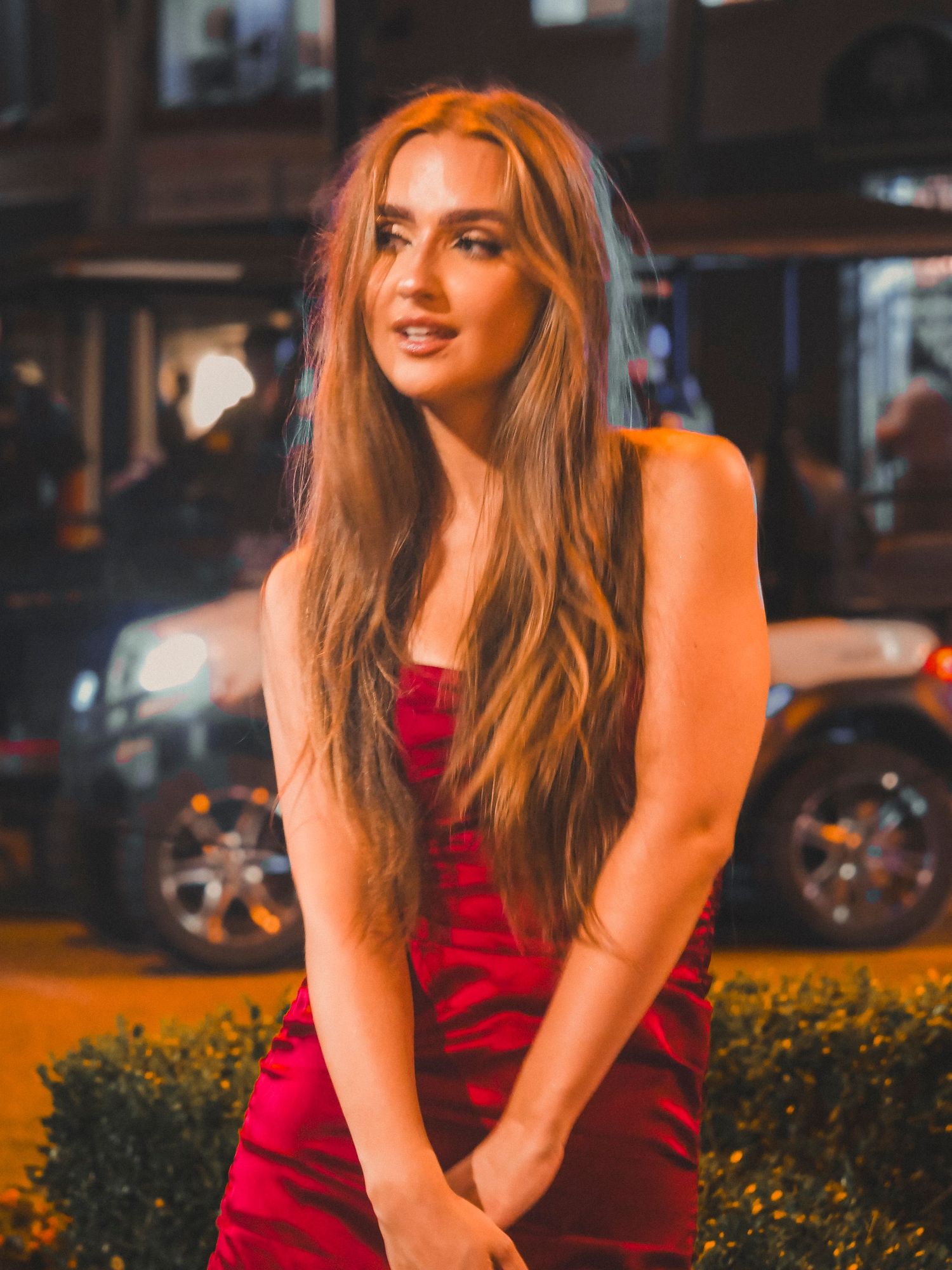 A woman in a red dress poses outdoors during golden hour lighting with urban architecture in the background.
