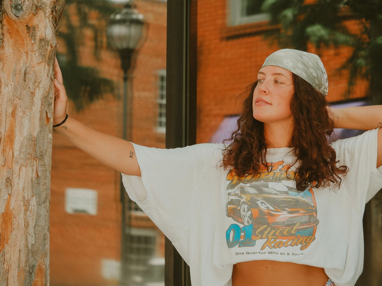 Young person in a white crop top and beanie poses with arms outstretched against orange brick building in warm sunlight.