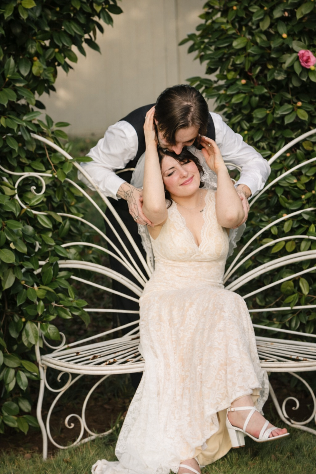 A figure in a white lace dress sits gracefully on a decorative white metal garden bench surrounded by lush greenery.