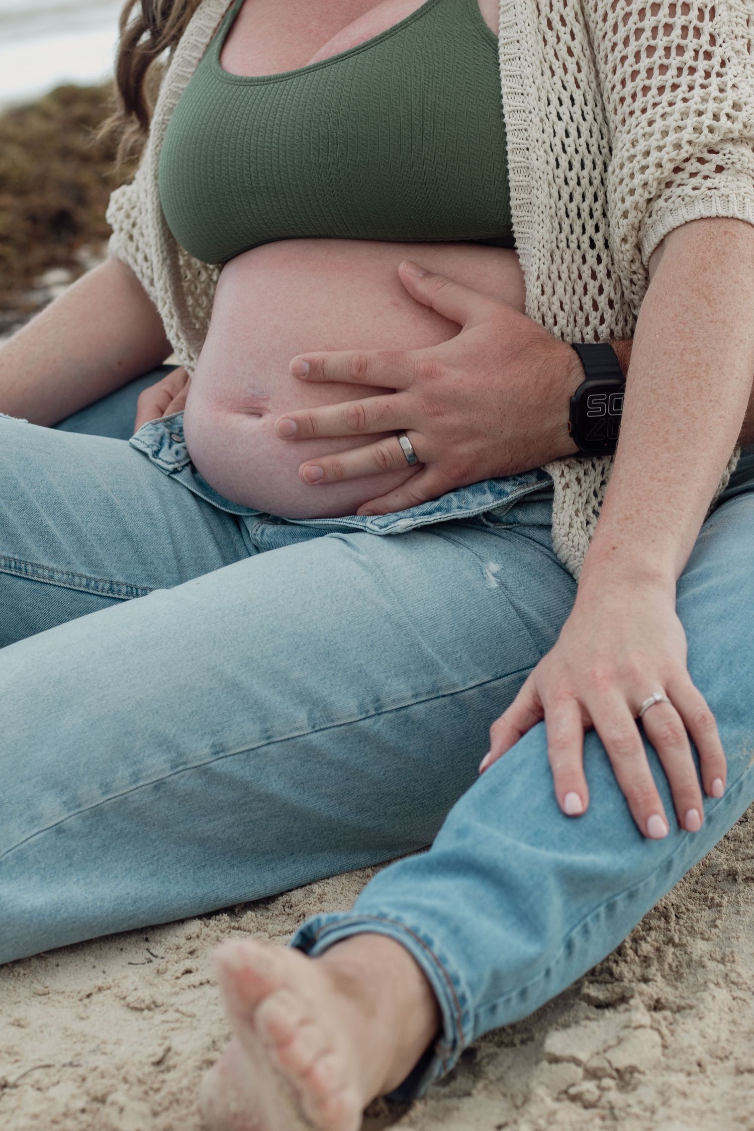 Close-up series of a pregnant woman in jeans and knit cardigan resting hands on her belly on a sandy surface.