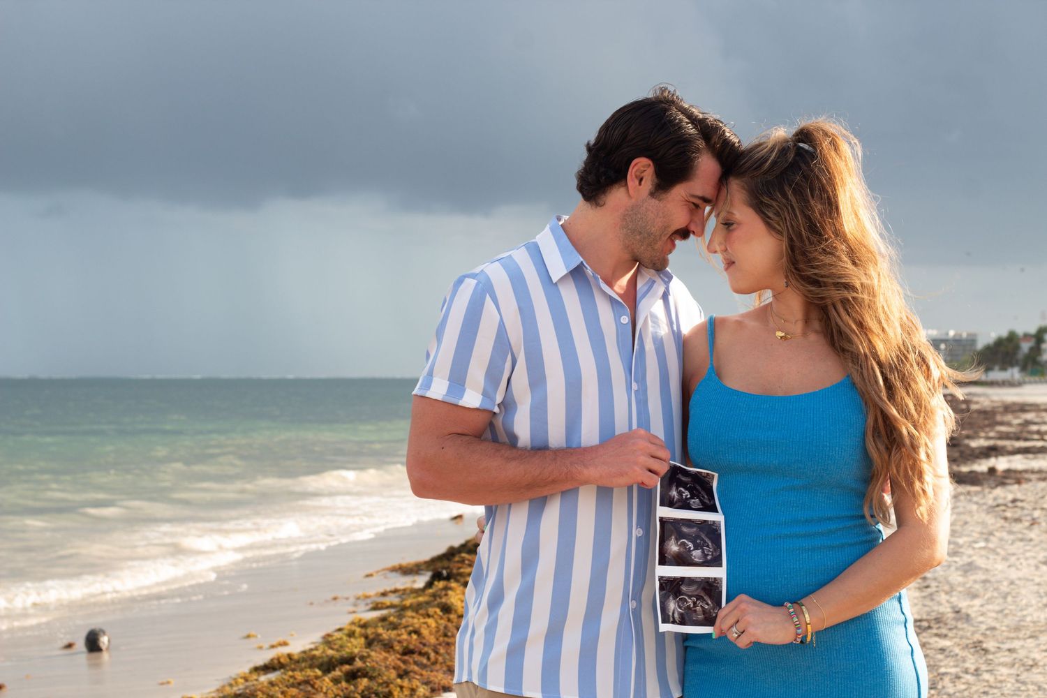 Couple sharing an intimate moment on a beach while holding an ultrasound photo by the ocean.