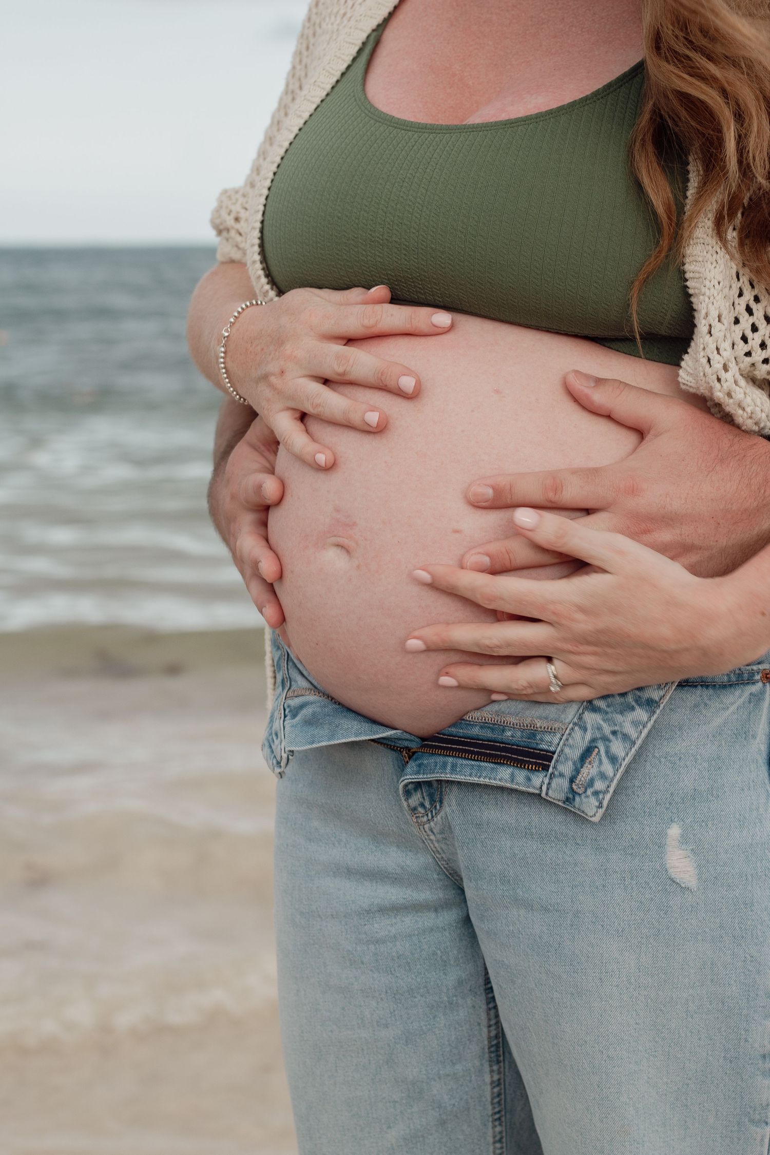 Close-up of pregnant belly at beach with hands cradling baby bump, wearing olive green top and distressed jeans.