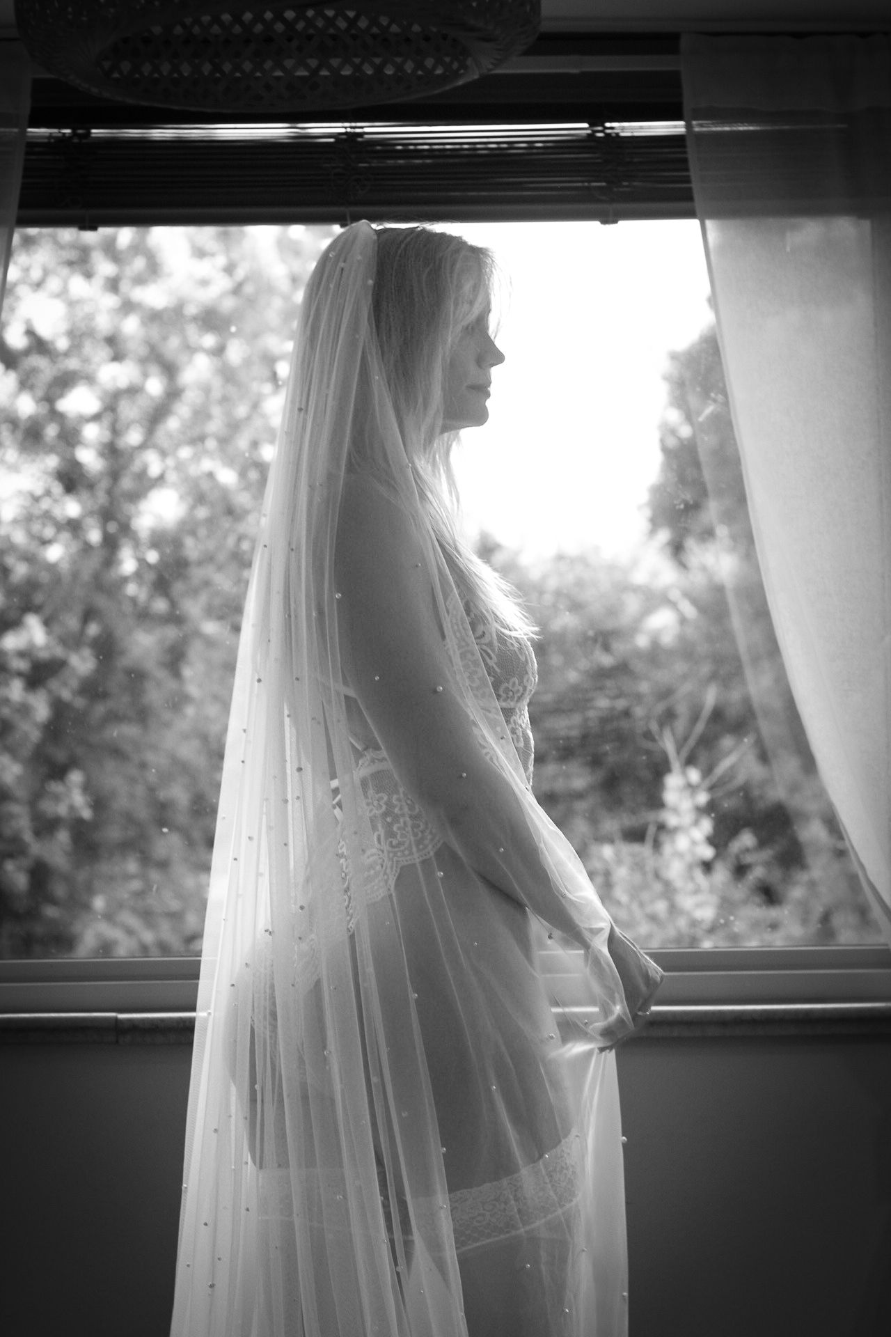 A bride in a flowing white veil stands silhouetted by window light in this elegant black and white photograph.