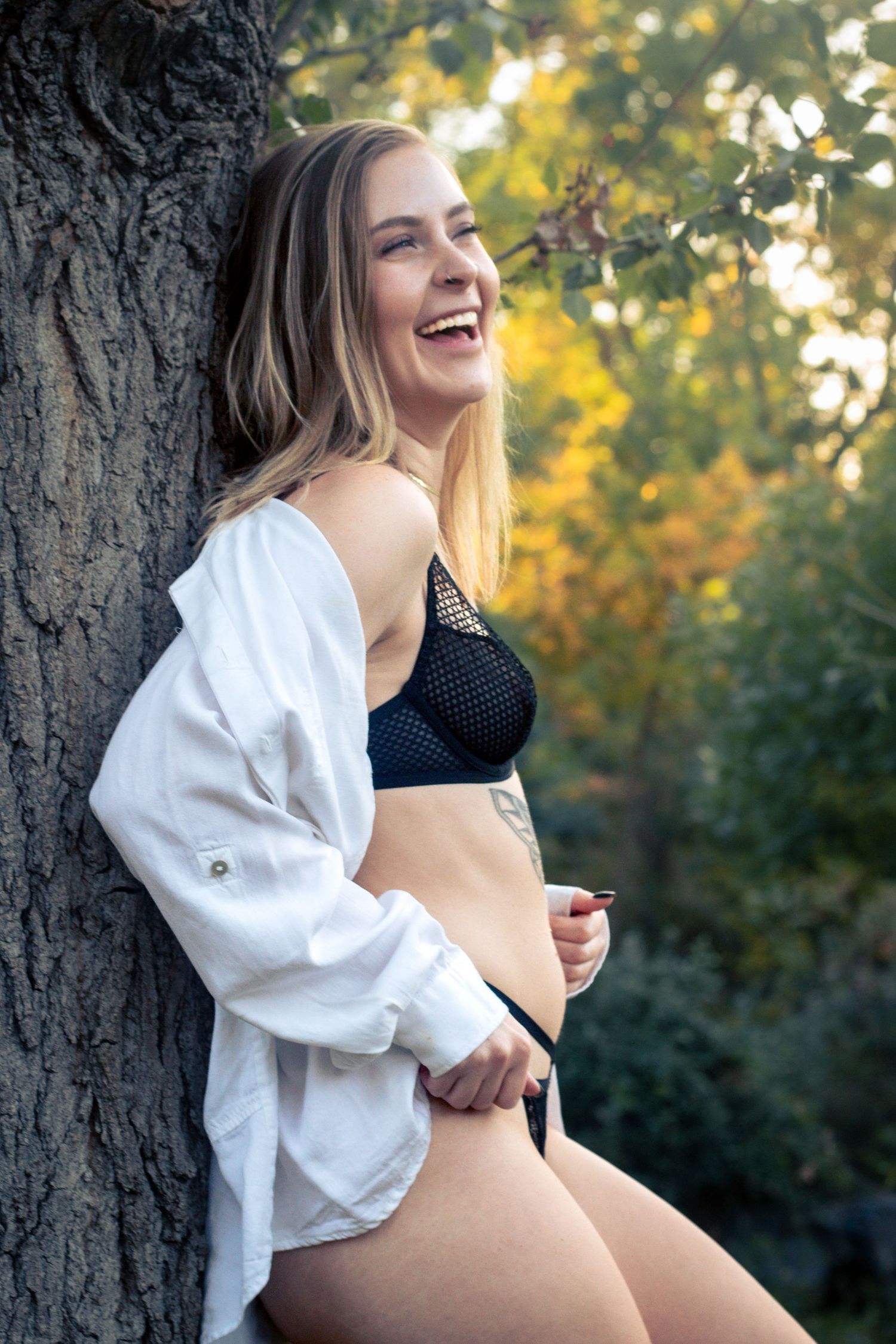 Lifestyle portrait of a joyful moment in black swimwear and white shirt leaning against a tree trunk with autumn colors behind.
