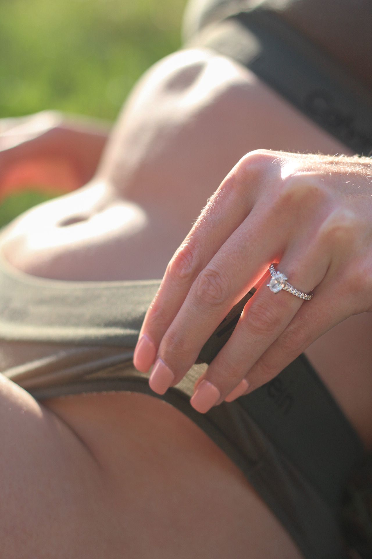 A delicate engagement ring sparkles in sunlight on a hand resting against dark fabric with soft bokeh background.