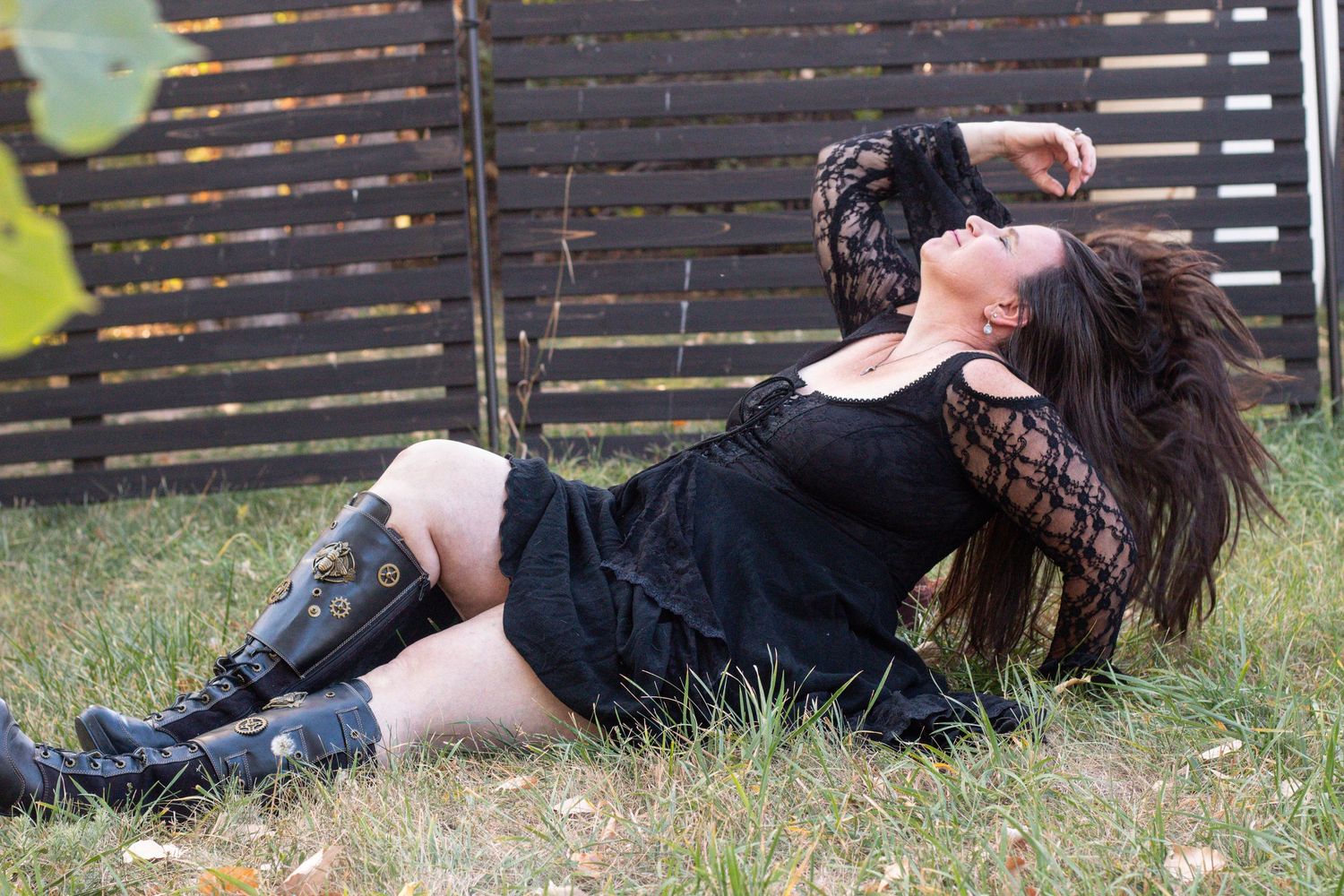 Figure in black dress and boots reclines on grass against wooden fence in outdoor portrait setting.