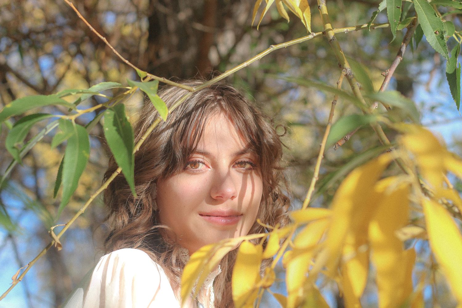 A girl in a white top poses among yellow autumn leaves and tree branches in soft sunlight.