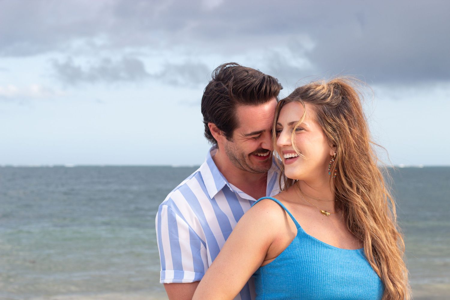 Romantic couple shares an intimate moment on a scenic beach with cloudy skies and ocean waves in the background.