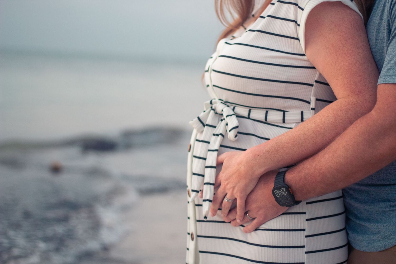 Expectant father holds expectant mother's belly while standing next to the ocean shore