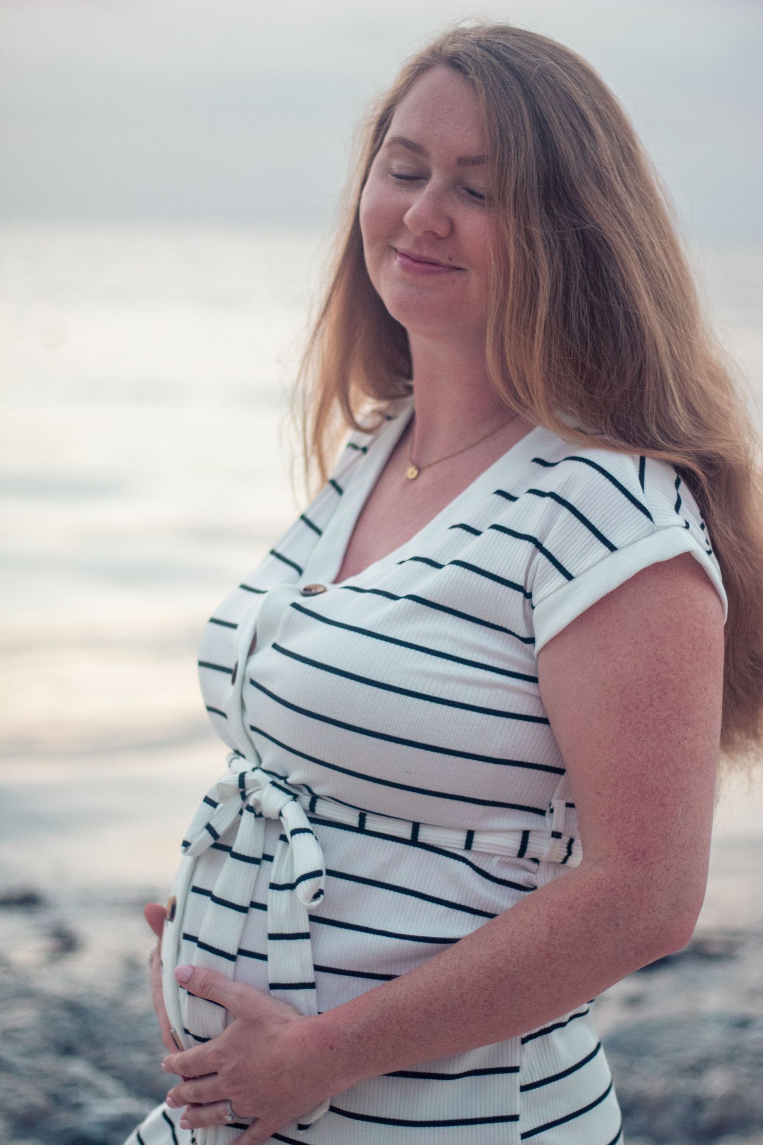 A pregnant woman in a striped white dress stands peacefully by the water at sunset.