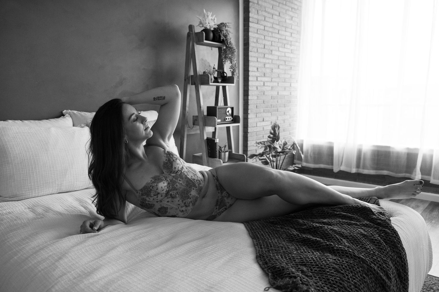 Black and white image shows person laying in bed with morning light coming through window, creating moody shadows.