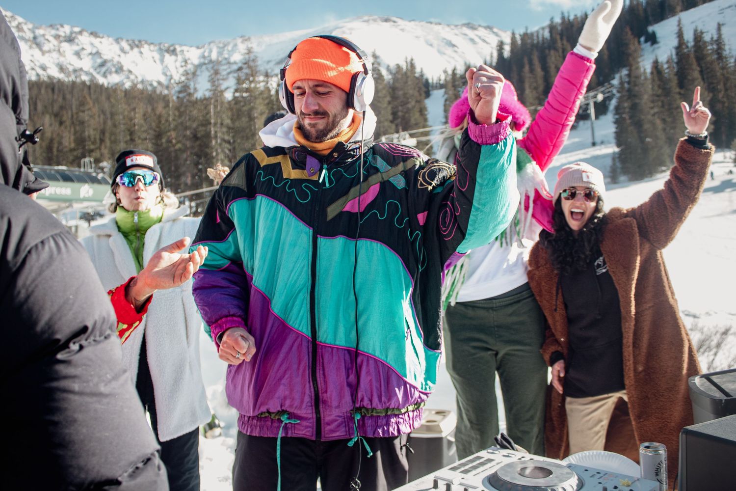 Skiers in retro bright ski jackets celebrate at Arapaho Basin base area with snow-capped peaks in the background.