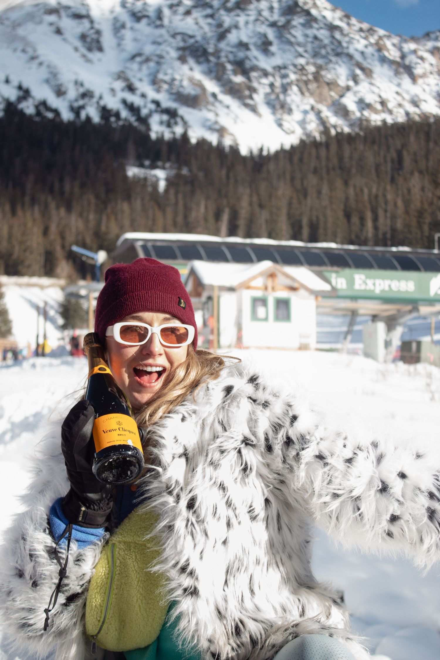 A person in a white fur jacket and ski goggles holds up a champagne bottle at Arapaho Basin ski area