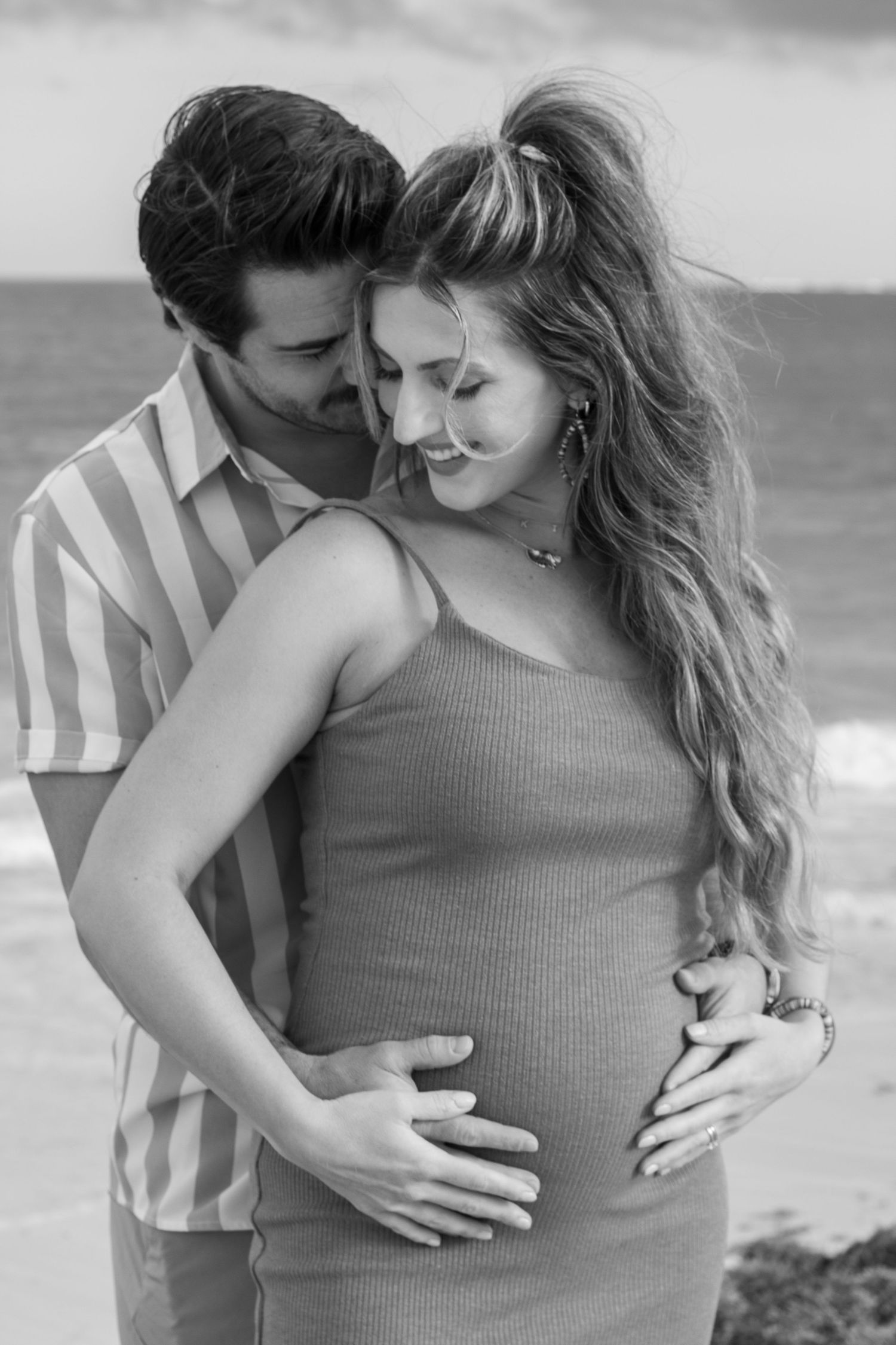 Black and white photo of a romantic moment between a couple at the beach, with a pregnant belly and gentle embrace.