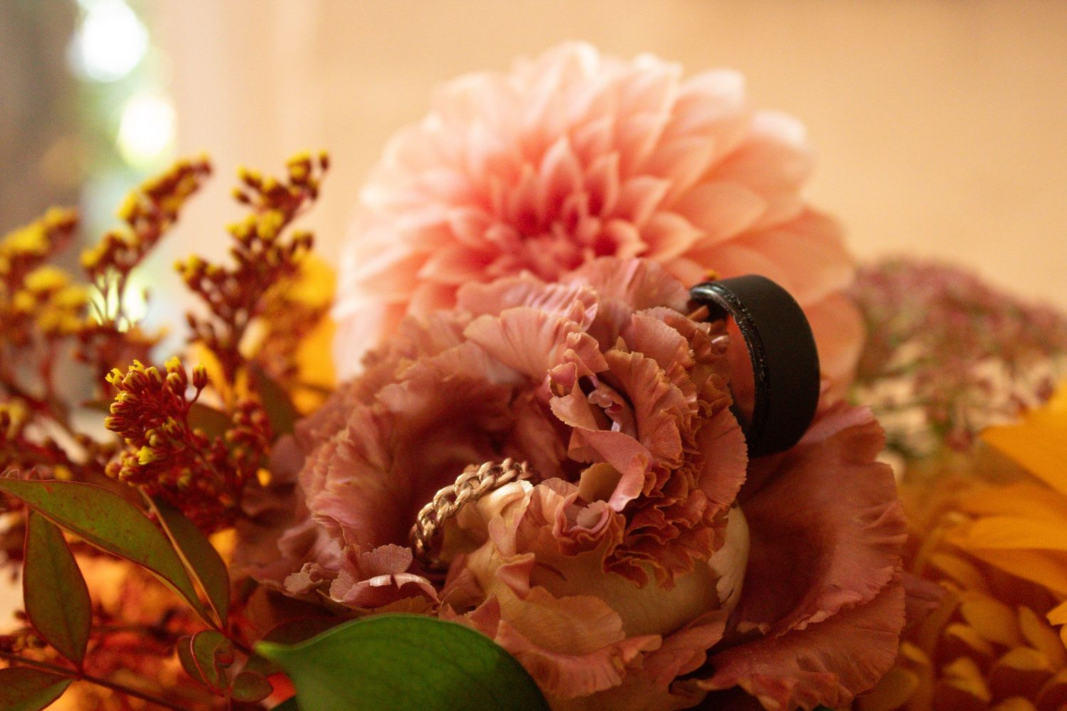 Close-up macro photography of wedding rings draped against pink carnation flowers with backlighting creating a warm, dreamy glow.