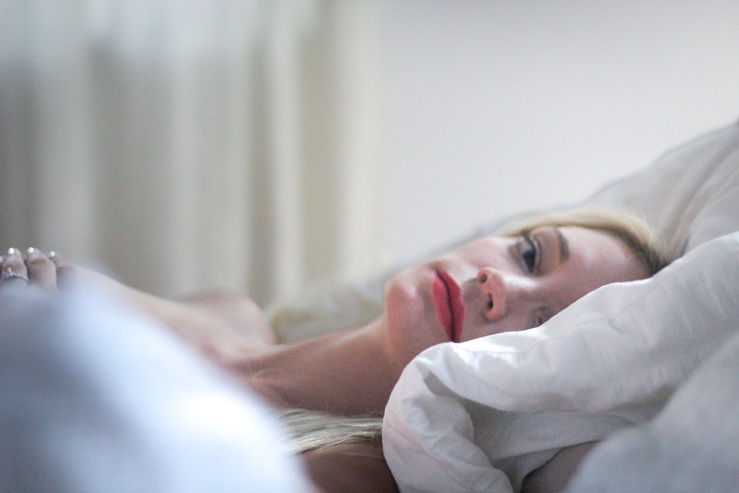 Person lying in bed with white bedding in soft morning light through window.