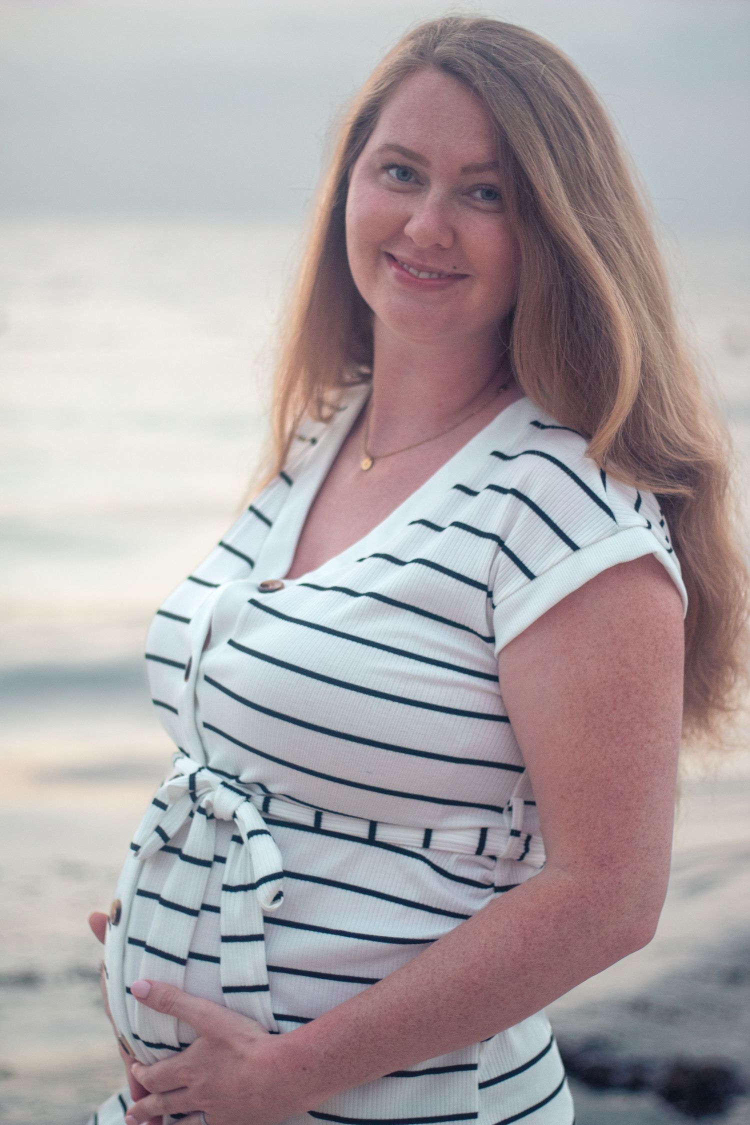 Woman wearing a white and navy striped dress poses at the beach during sunset, showing off her pregnant belly