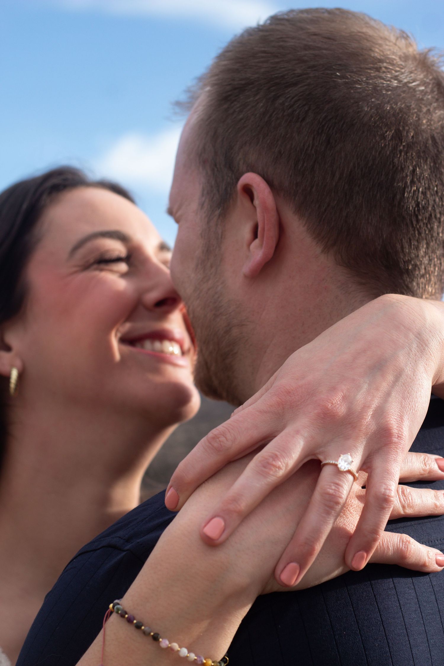 Close up of hands showing an engagement ring during a romantic outdoor moment against a blue sky.