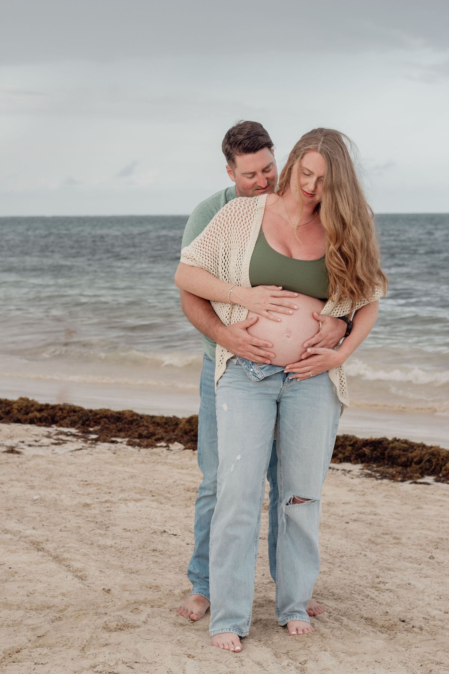 Expecting couple shares intimate moment on beach during maternity photoshoot with ocean backdrop.