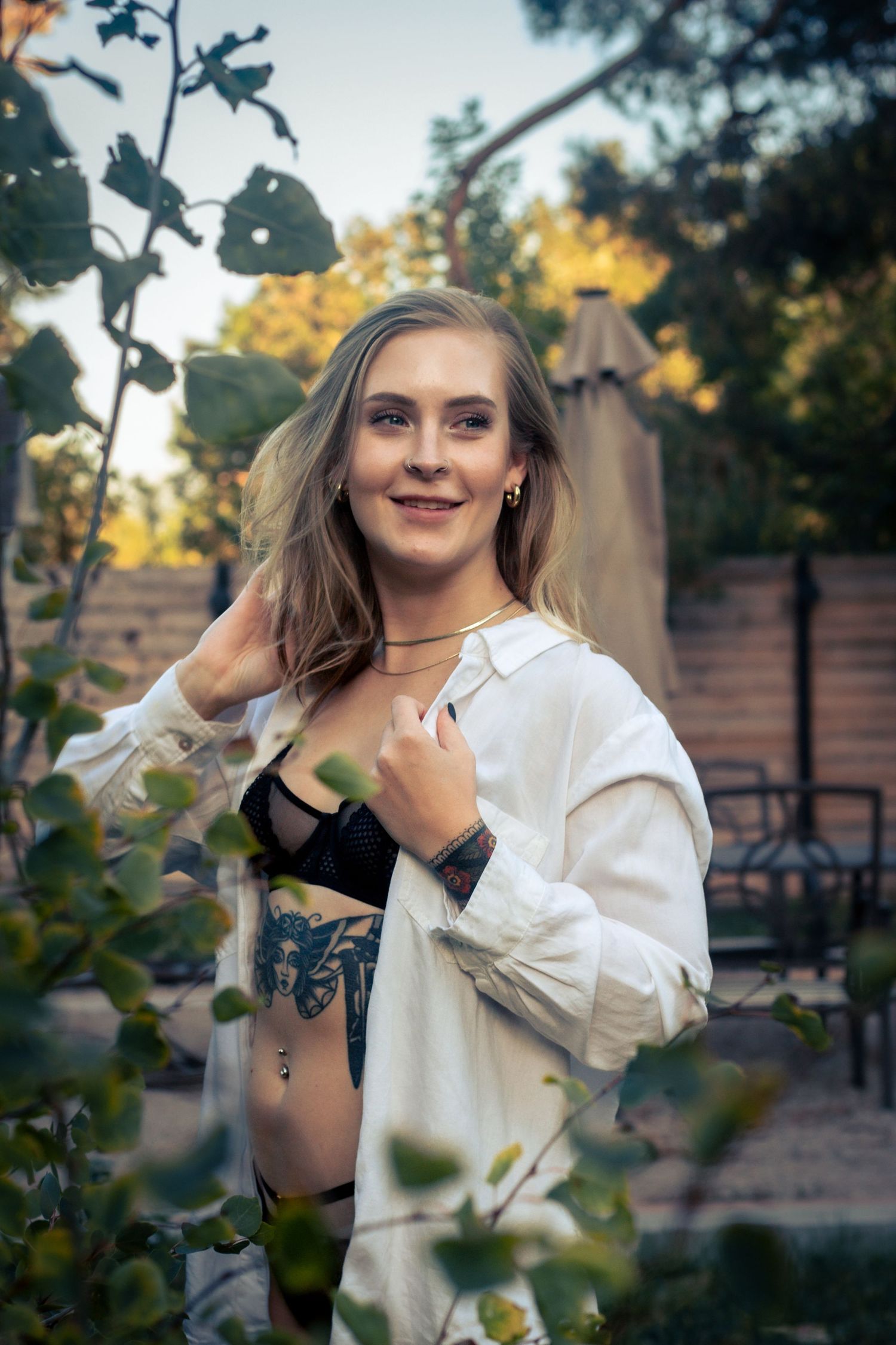A person wearing an open white button-down shirt and black top poses outdoors near foliage at sunset.