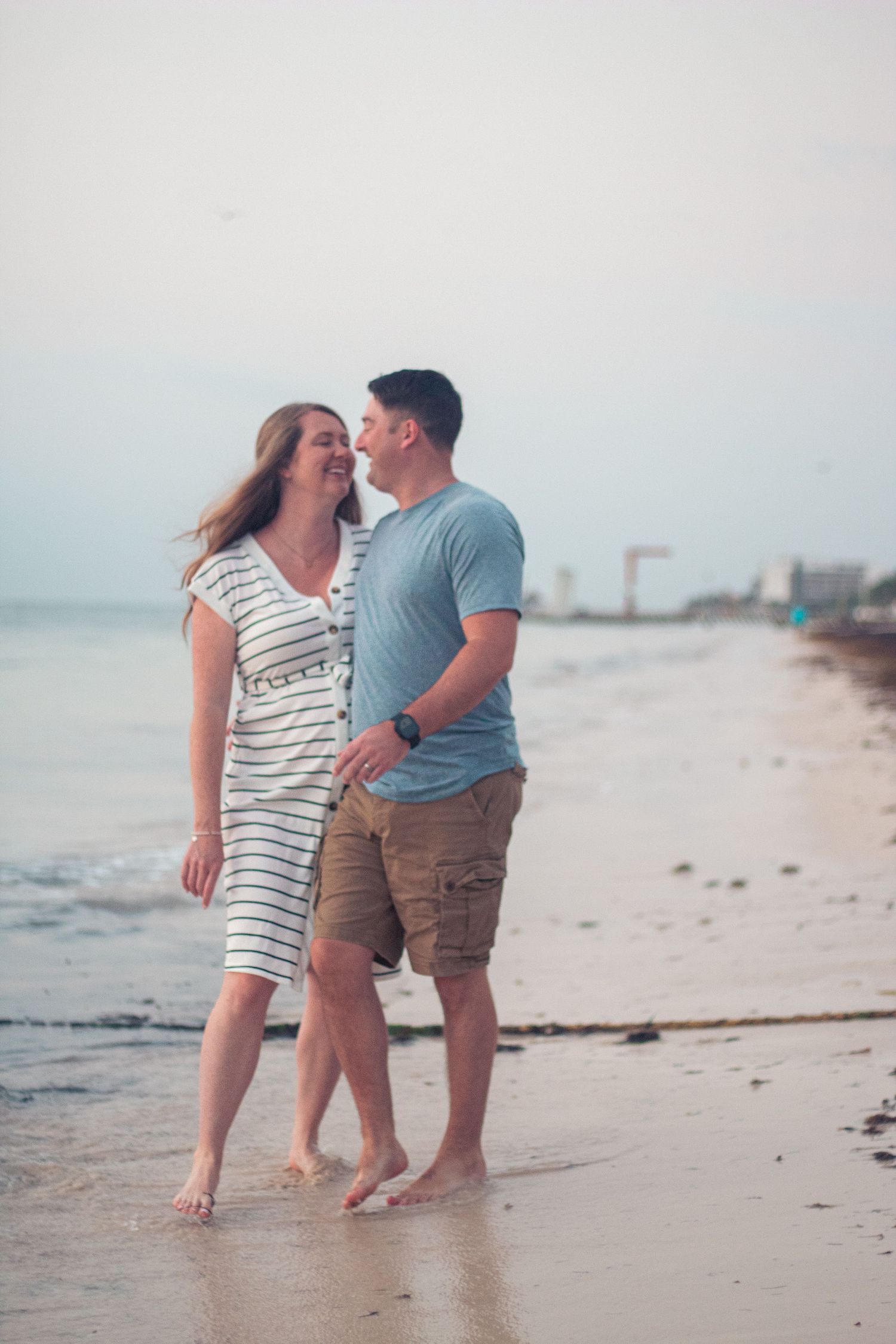 A romantic couple stands barefoot on a beach at sunset sharing an intimate moment together.