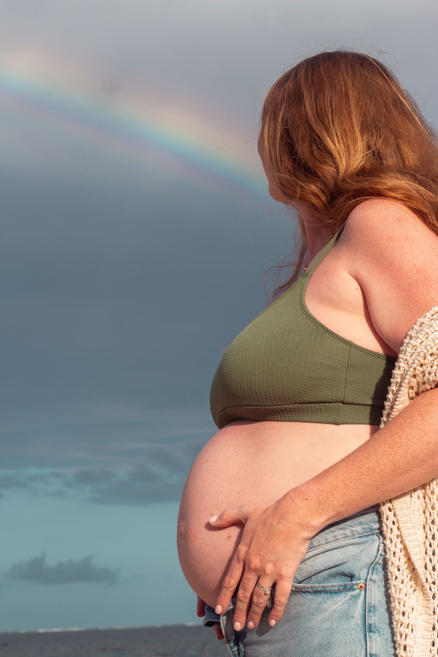 A pregnant woman in a green crop top and jeans stands against a cloudy sky with a rainbow in the background.
