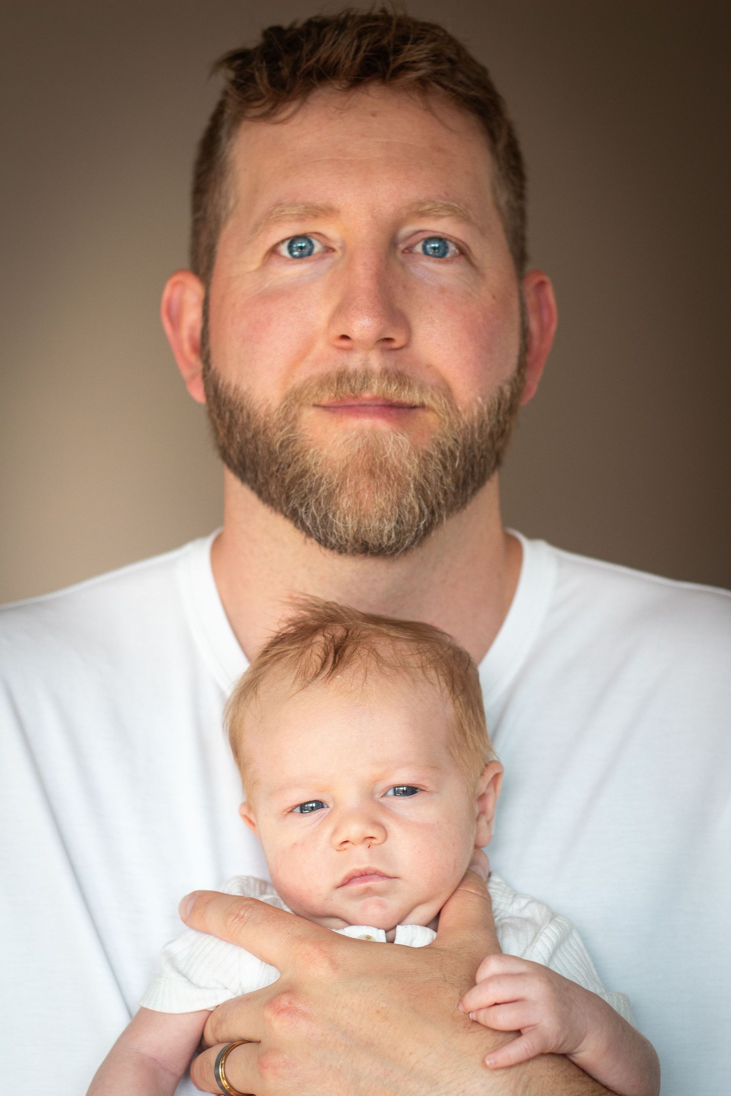 Father holds infant son in front of him while showing off their matching blue eyes in front of a beige background