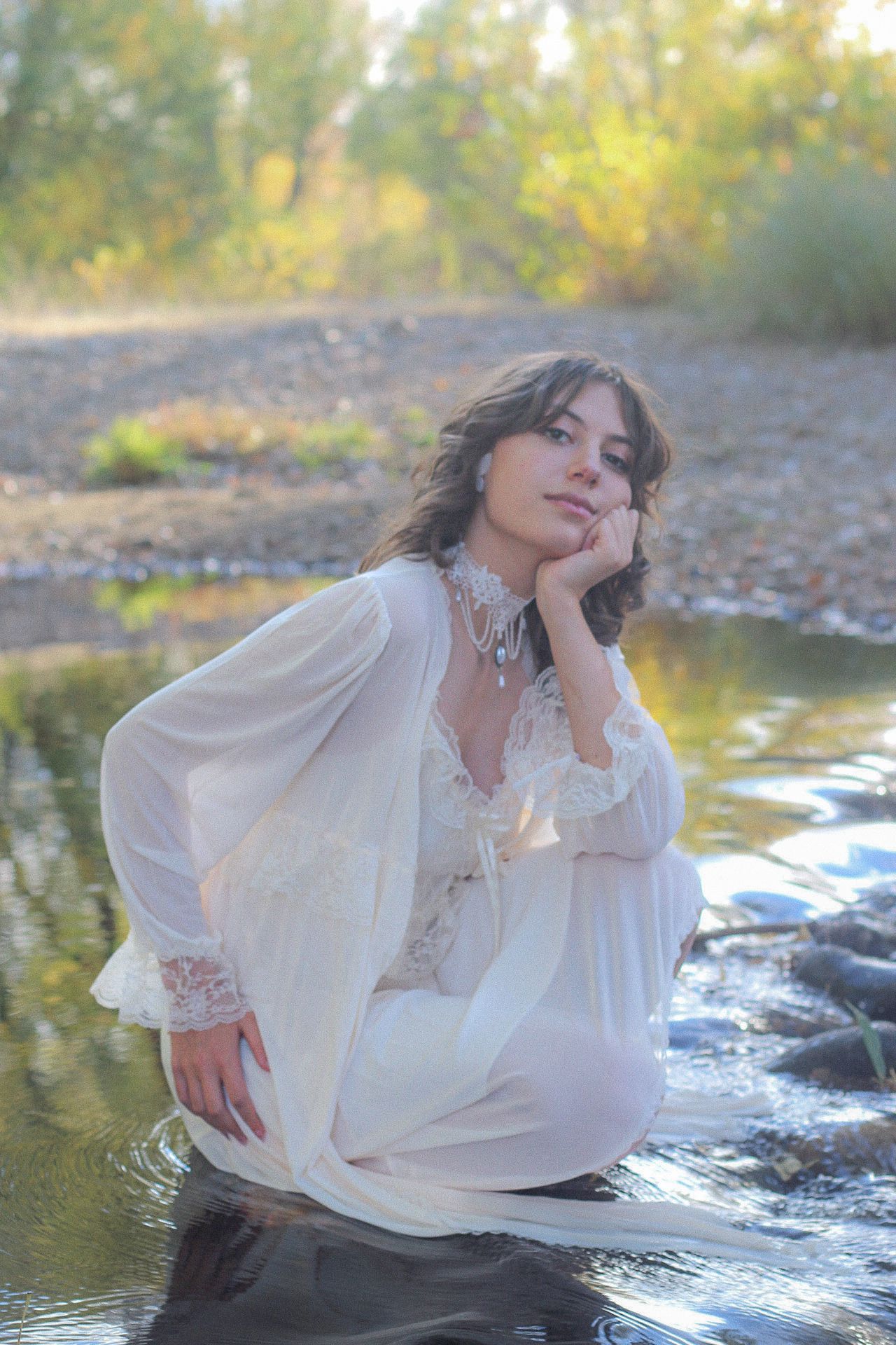 A girl in a flowing white dress poses thoughtfully by a stream in soft natural lighting during golden hour.