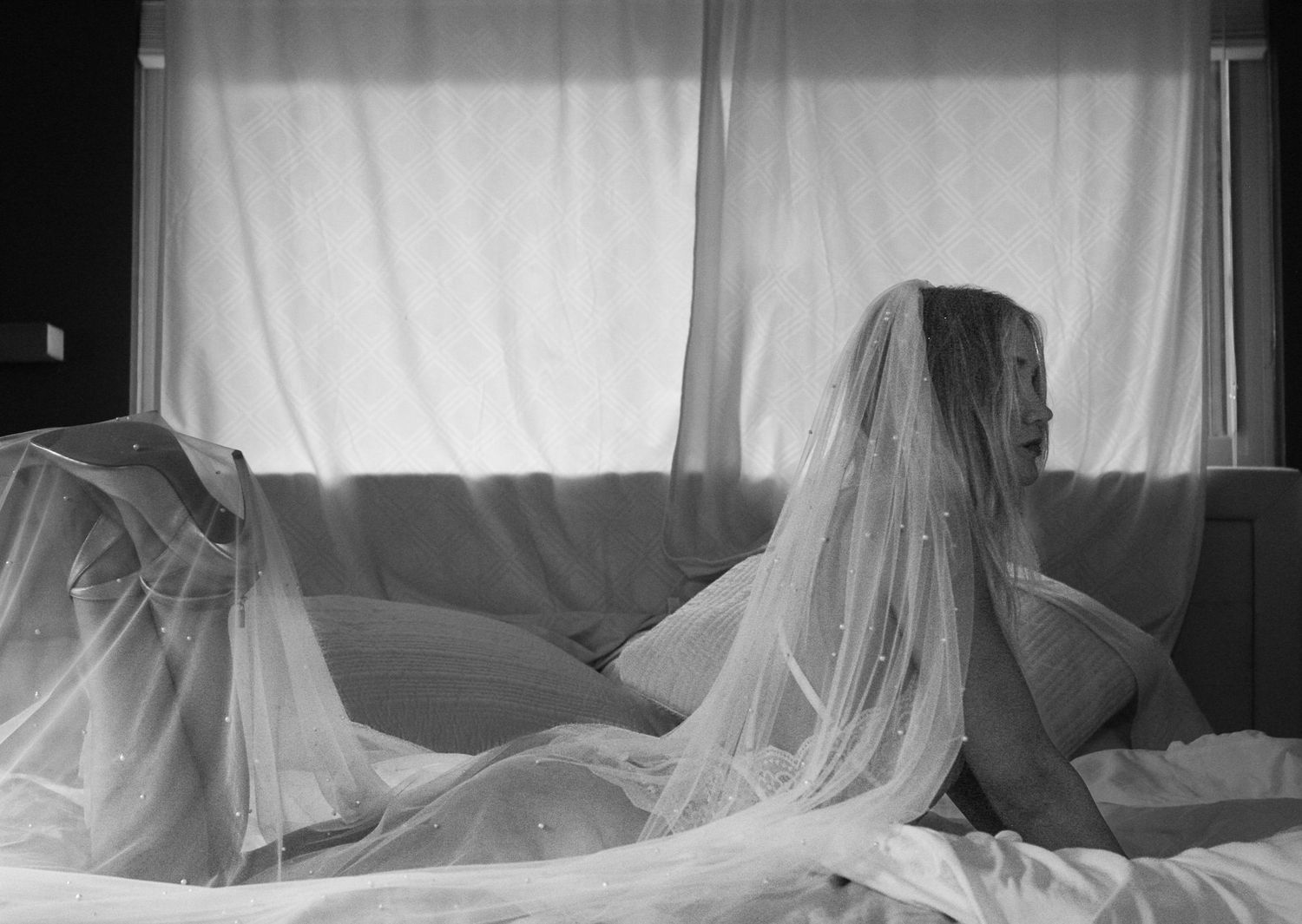 A figure draped in flowing white veil fabric sits on a couch in black and white photography.