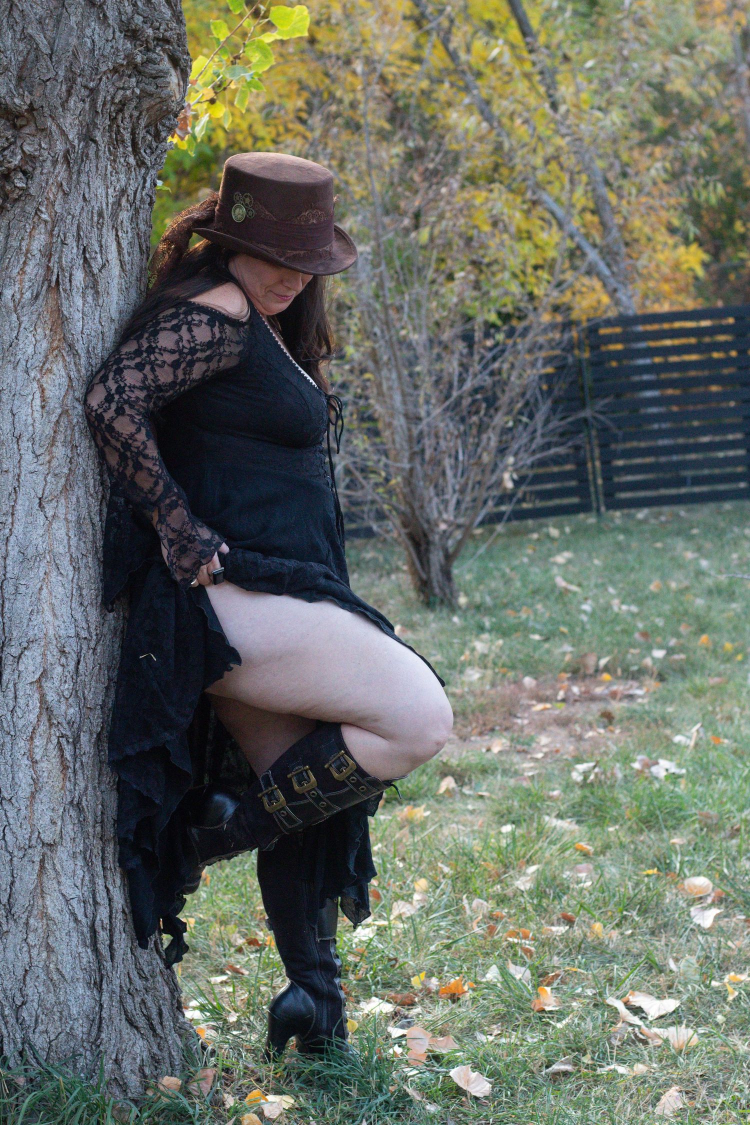 A person in a steampunk-inspired outfit with a brown hat, lace sleeves and boots poses against a tree in autumn.
