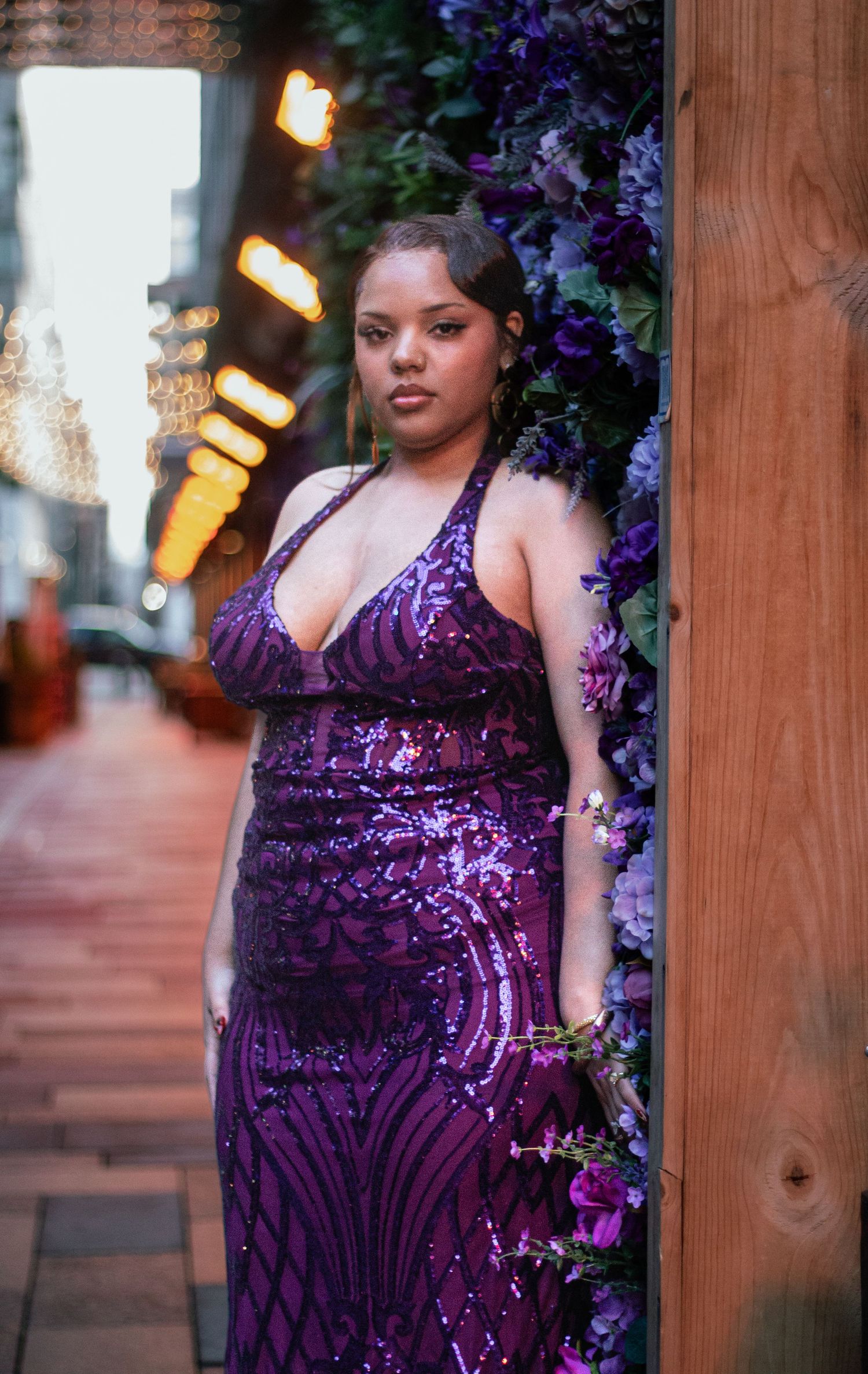 A woman going to prom in a sparkling purple sequined gown poses against wooden posts with purple flowers in view.