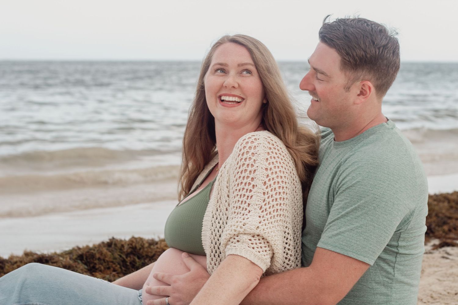 A couple sits together laughing on a beach with waves and seaweed visible in the background during maternity photoshoot