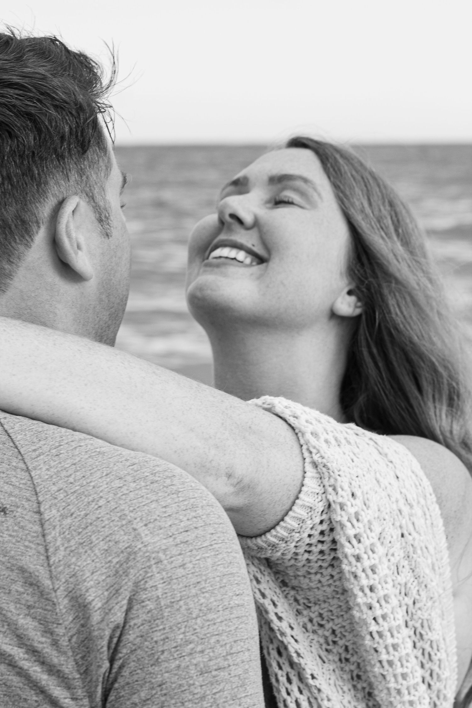 A romantic black and white photo of a couple laughing together by the ocean, wearing cozy sweaters.