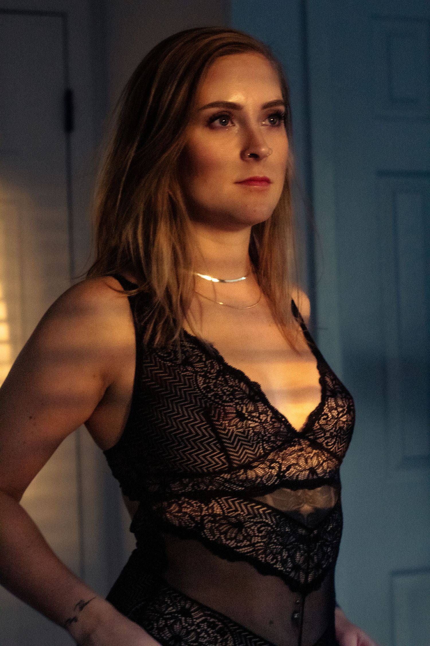 Woman in black lace lingerie poses in moody lighting against a wall.