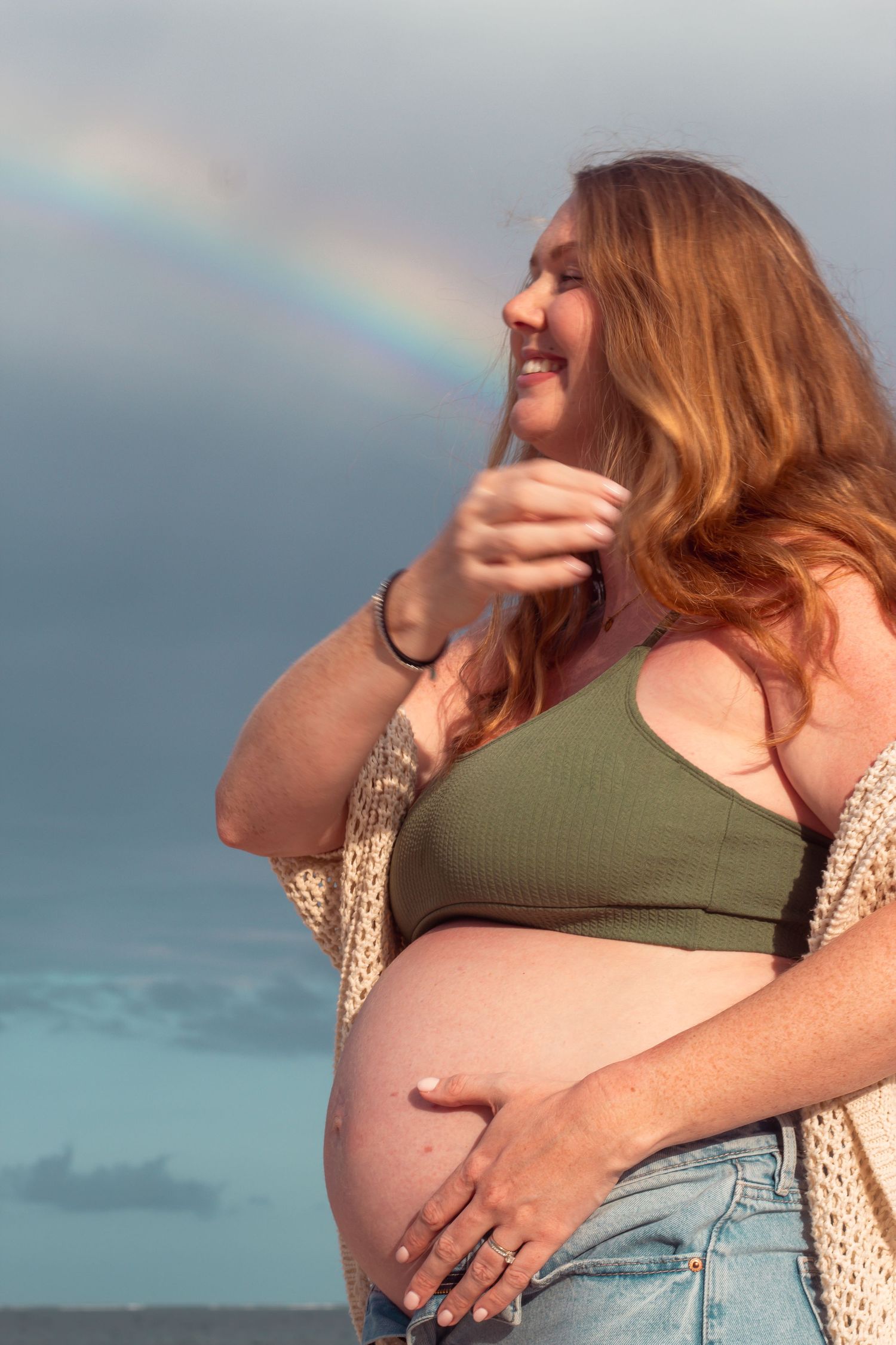 A pregnant woman in a green top and cardigan laughs joyfully as a rainbow appears in the cloudy sky behind her pregnant belly.