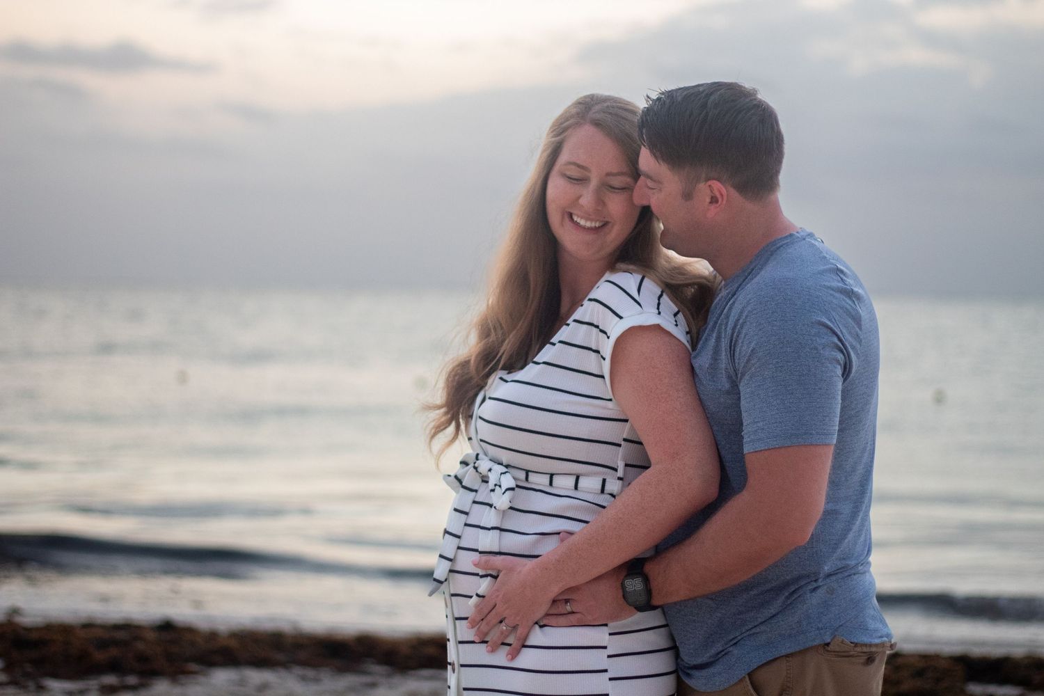 A couple embraces on a beach at sunset, with the pregnant woman wearing a striped dress and laughing joyfully.