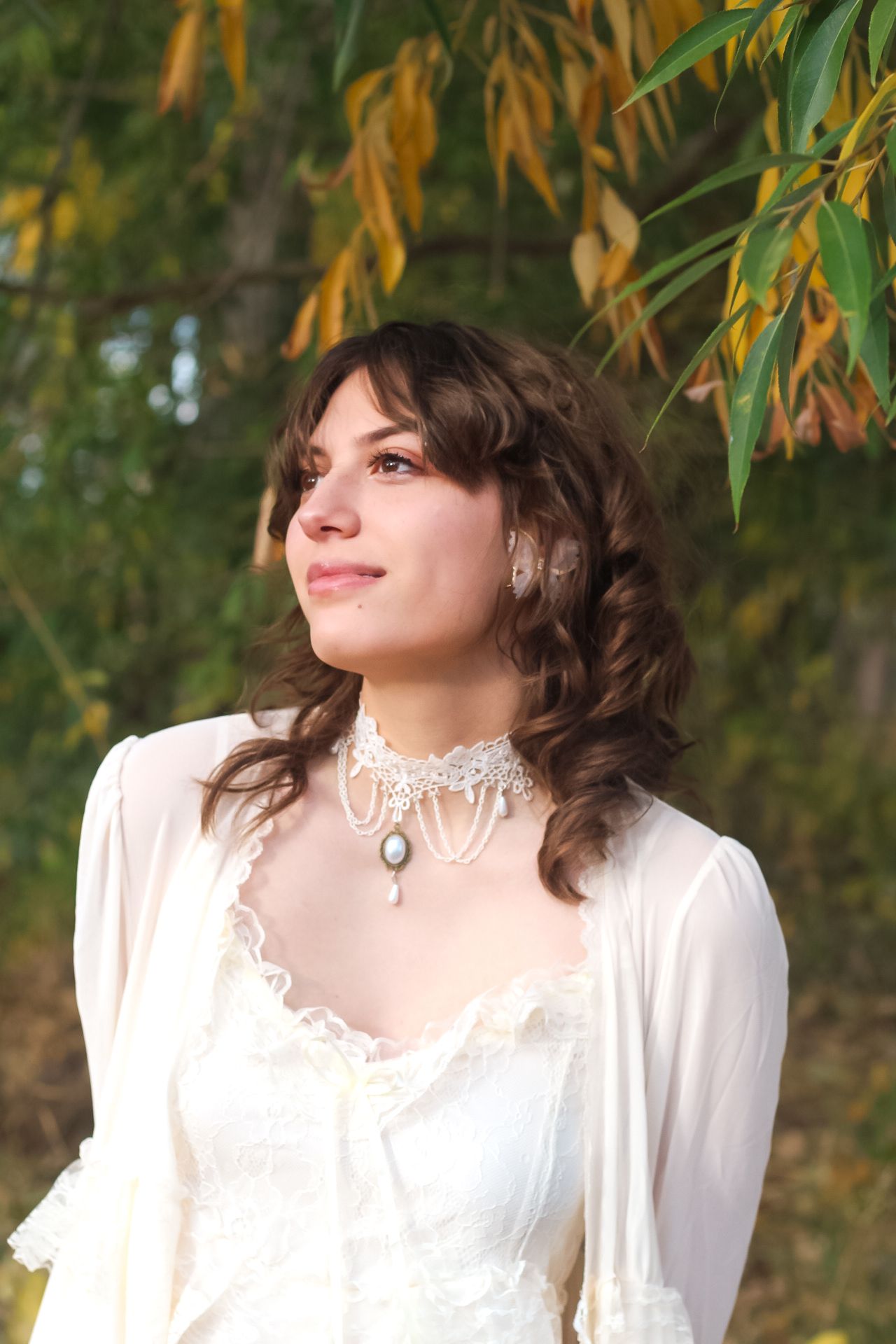 A dreamy outdoor portrait in white lace dress with vintage choker necklace against autumn foliage backdrop.