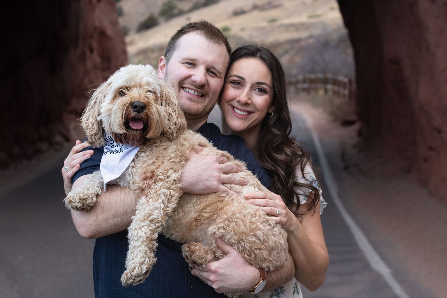 Two people and their goldendoodle puppy pose together outdoors for engagement photos at Red Rocks Amphitheater