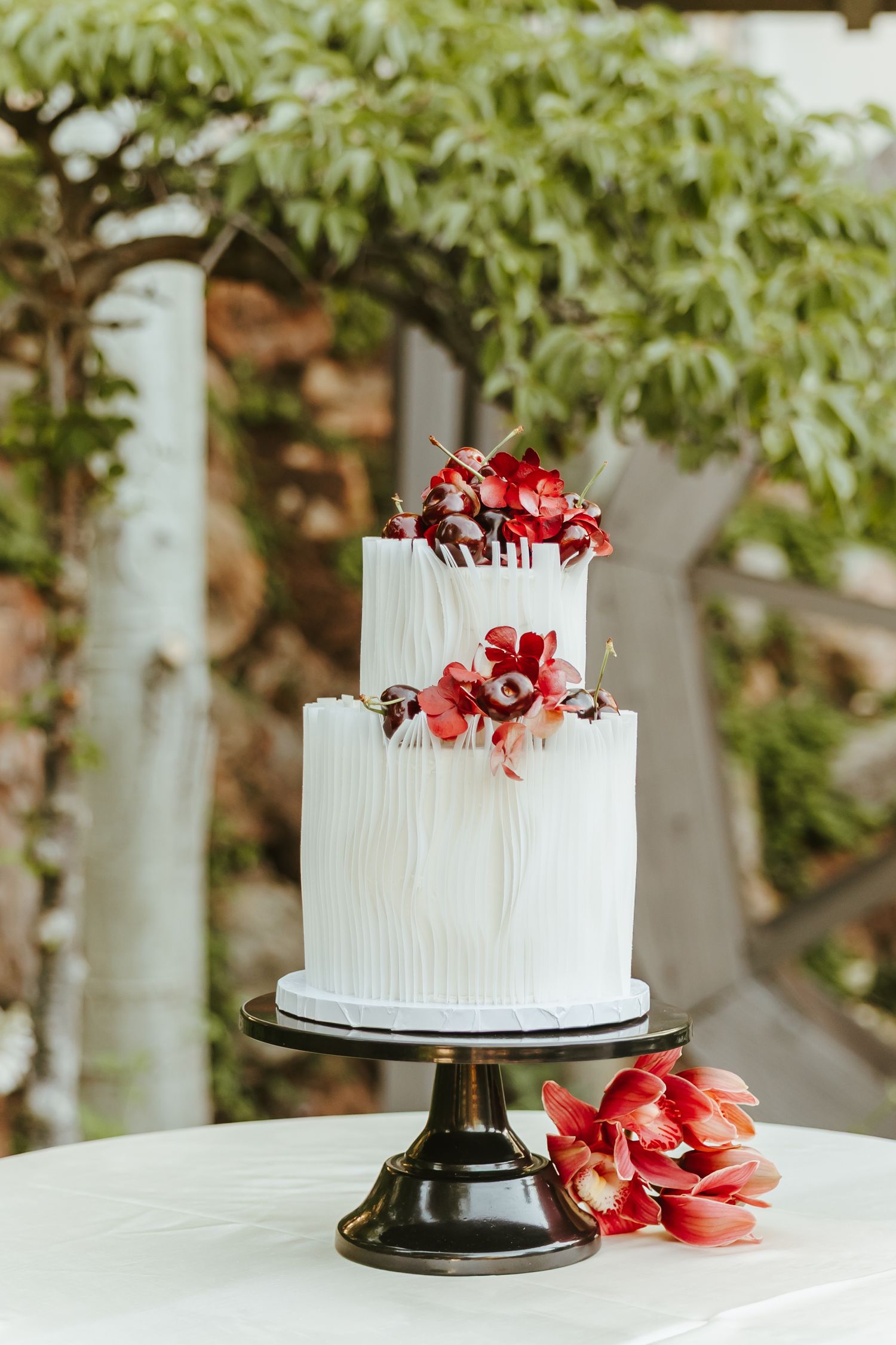 Elegant two-tier white wedding cake decorated with red orchids on black pedestal stand against garden backdrop.