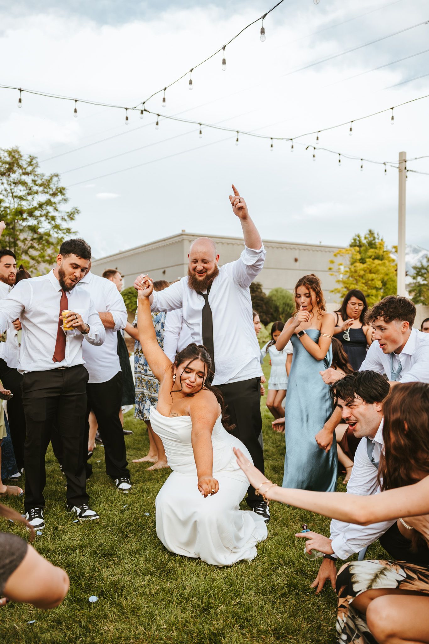 Wedding party dances and celebrates outside on grass lawn under string lights during lively outdoor reception celebration.