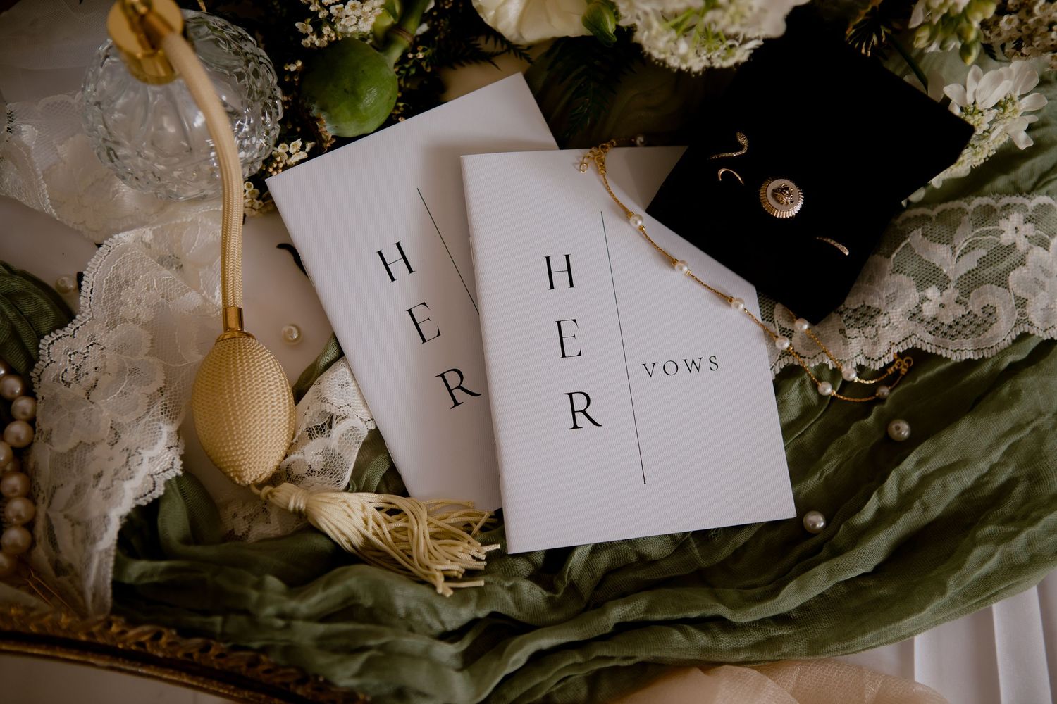 Wedding vows booklets labeled 'Her' rest among delicate lace and greenery with a wooden spoon decoration.