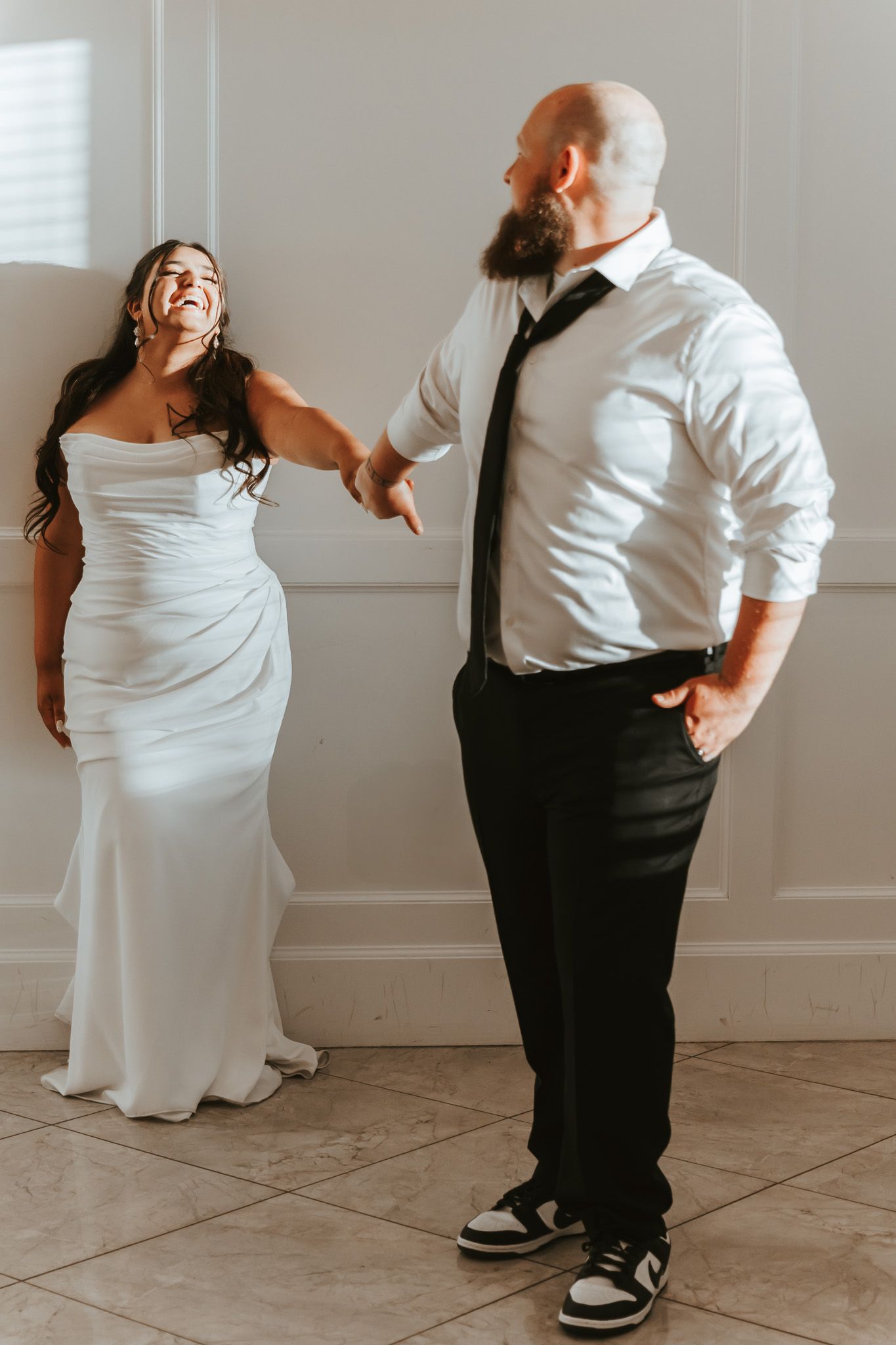 A bride in a white fitted gown shares a laughing moment with someone in a white shirt and black tie during a wedding.