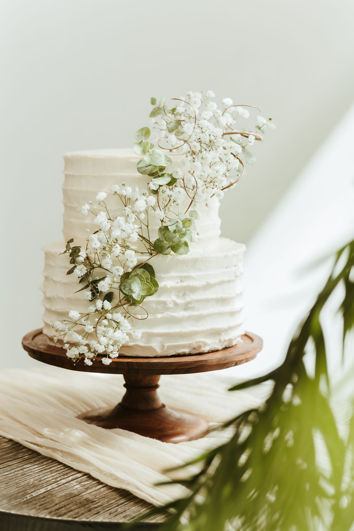 White two-tier wedding cake with textured buttercream frosting and delicate white floral cascade on wooden cake stand.
