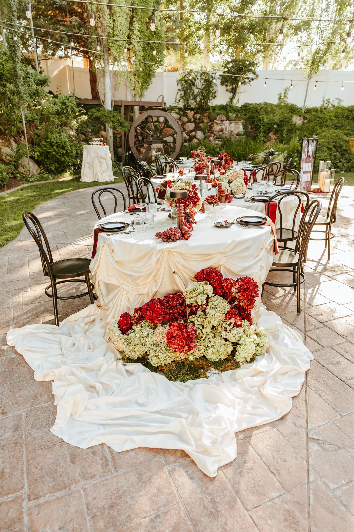 Elegant wedding reception table decorations featuring cascading red and white flowers on flowing white linens at an outdoor venue.