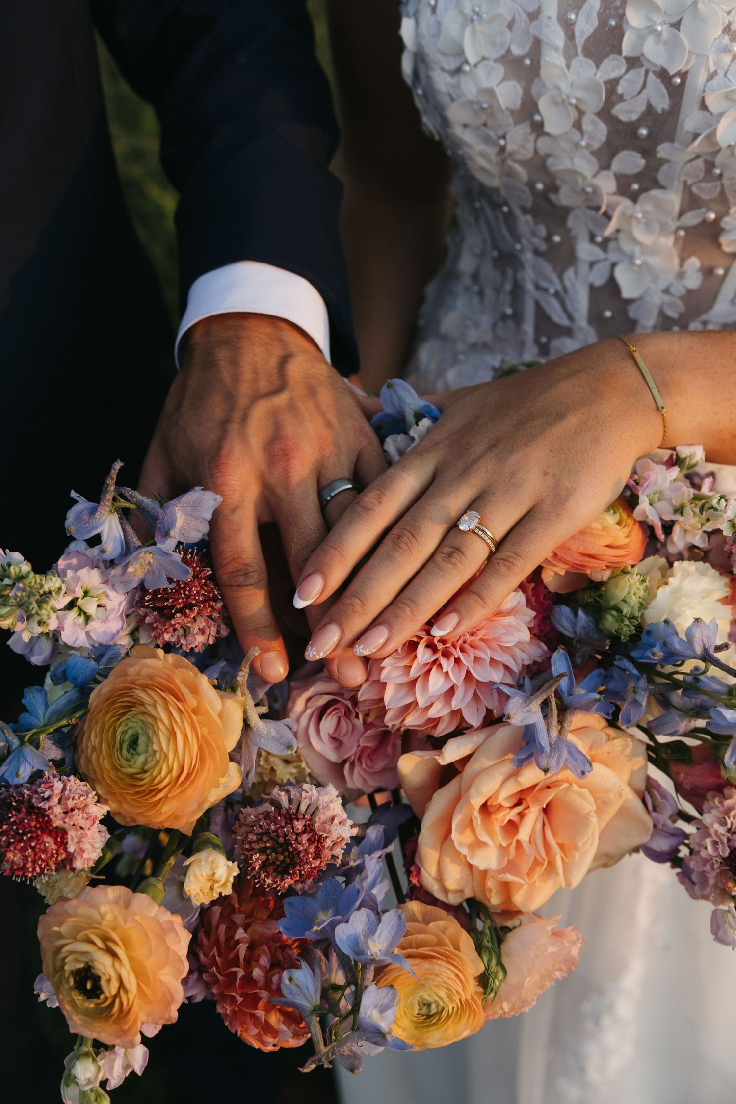 Hands with wedding rings rest on a vibrant bridal bouquet with peach ranunculus, pink dahlias and blue delphinium flowers.