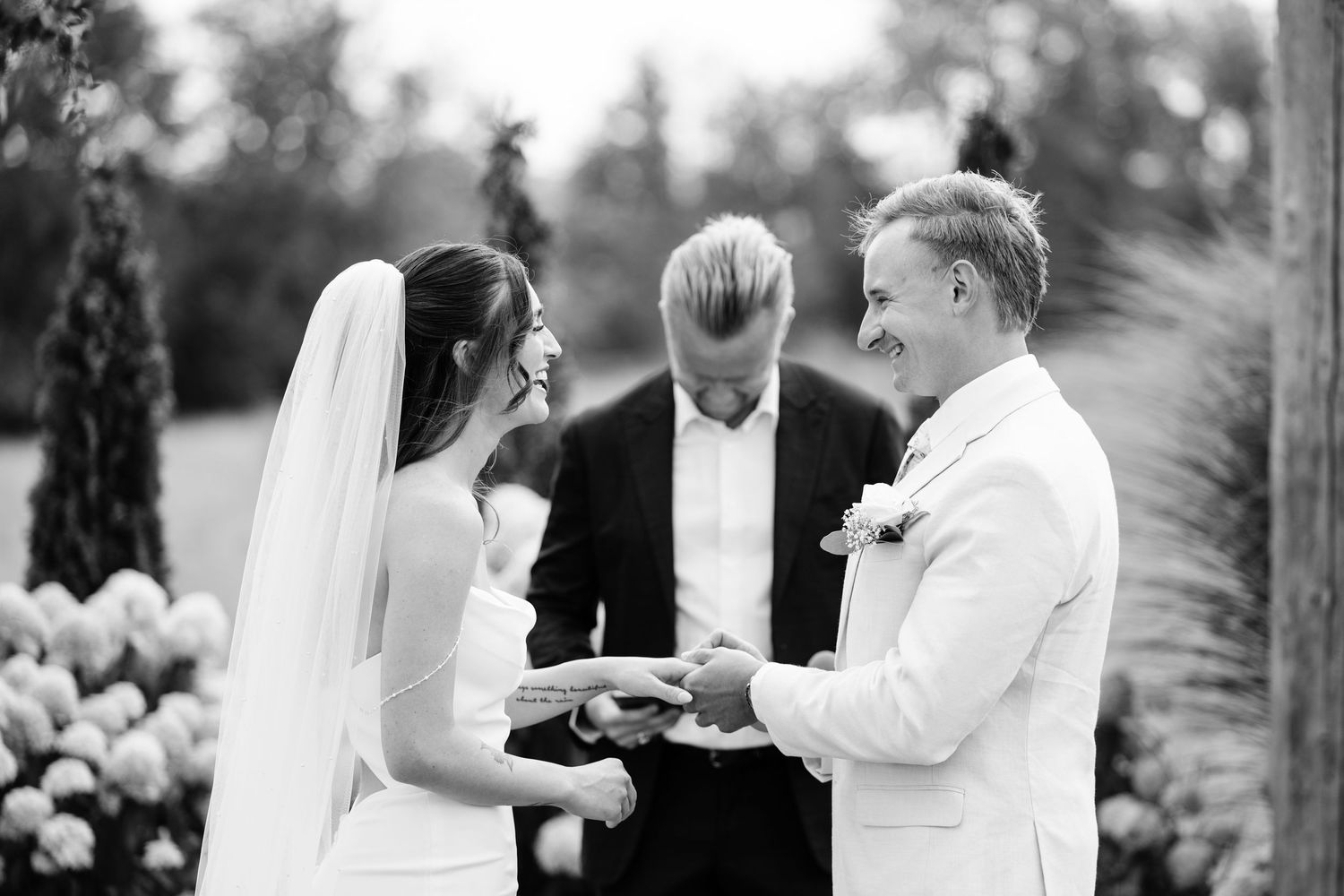 Black and white wedding ceremony photo of bride and groom exchanging rings outdoors with officiant.