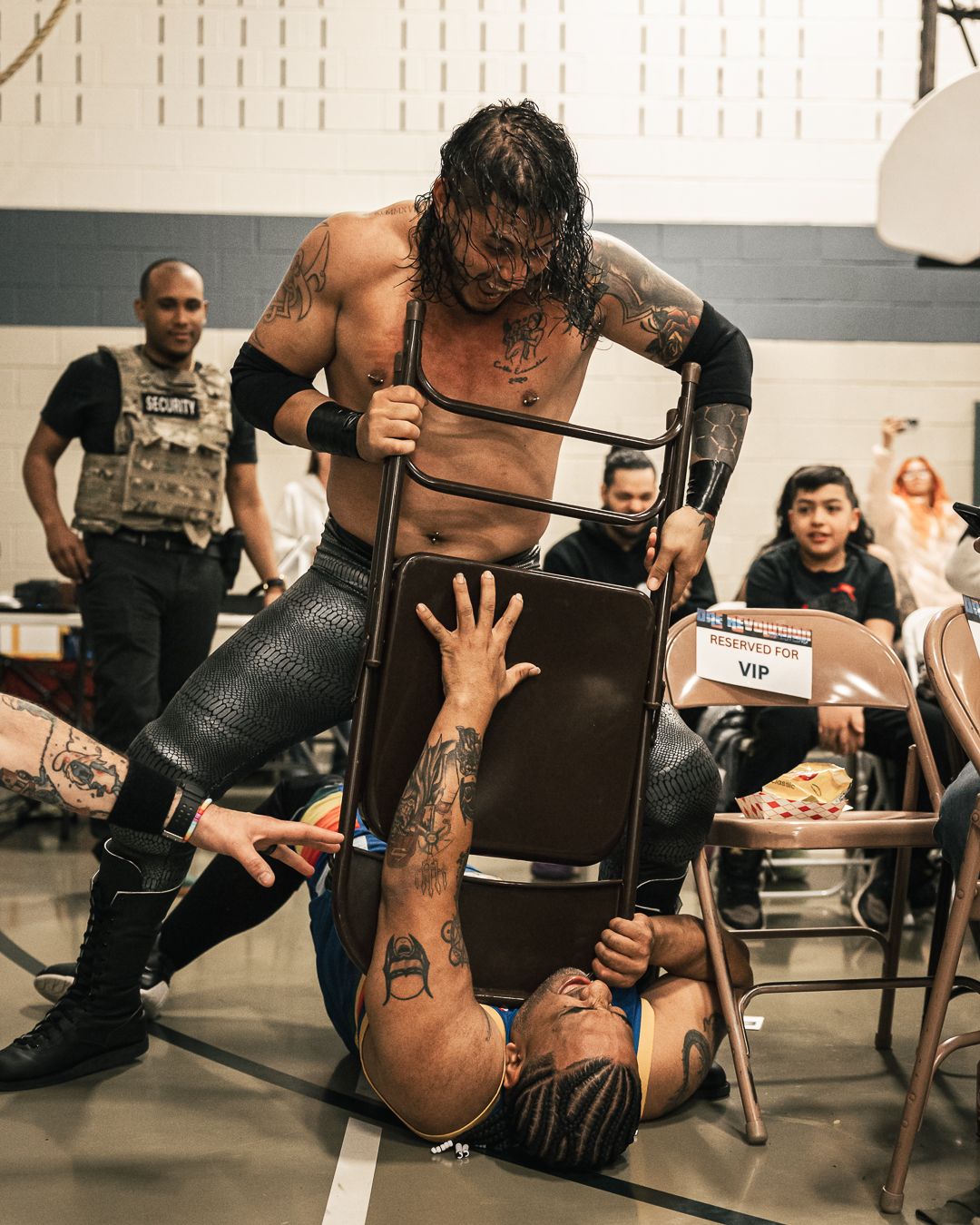 Two wrestlers perform a chair spot at an indie wrestling event, with one standing over a downed opponent holding a metal folding chair.